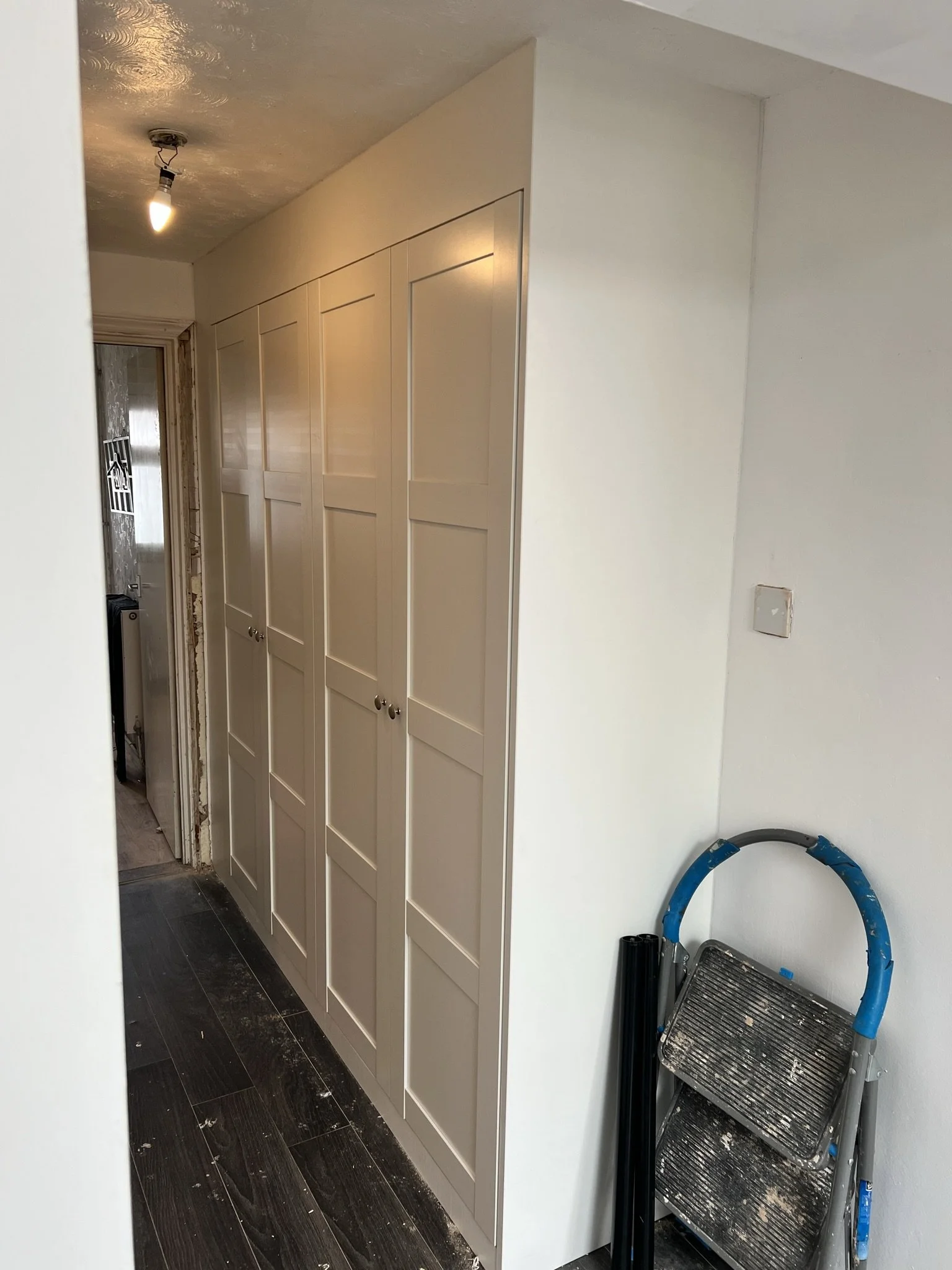 A hallway with beige paneled cabinets on the right, a ceiling light, dark wood flooring, and a folded step ladder leaning against the wall.