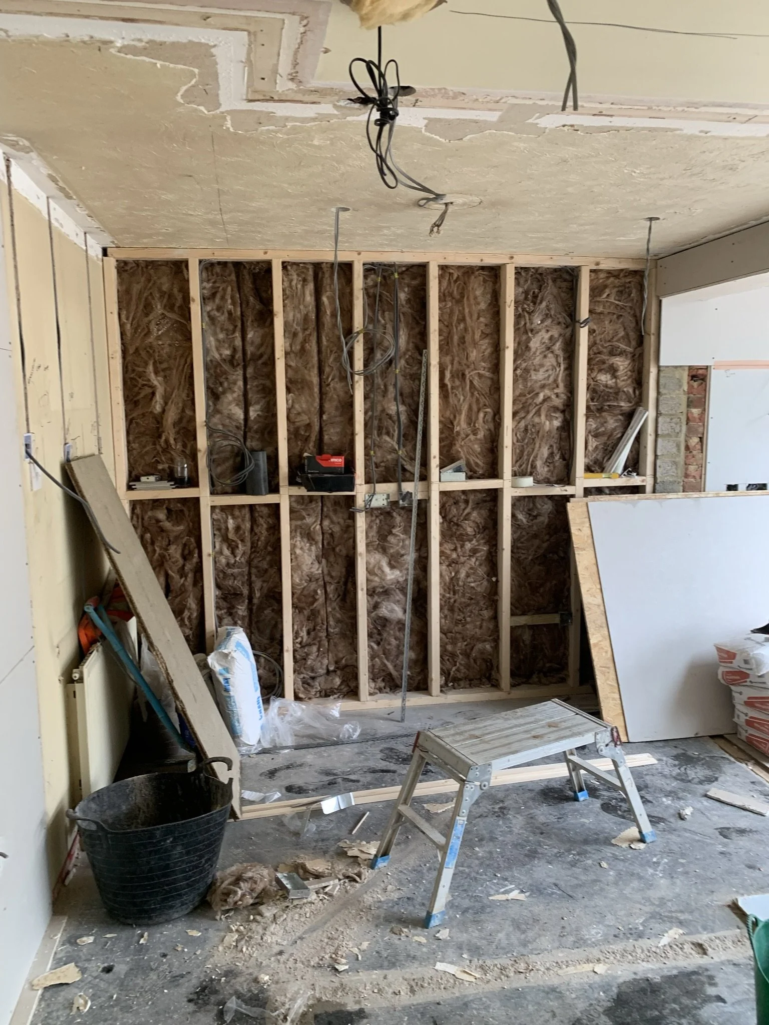 Room under renovation with exposed wall studs, insulation, and construction materials scattered on the floor. A metal step stool is placed near the center and electrical wires hang from the ceiling.