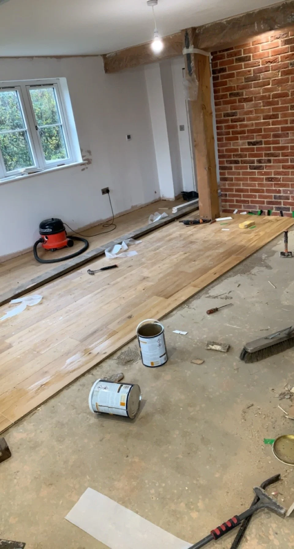 Room undergoing renovation with wooden flooring partially installed, tools scattered, a brick accent wall, and an upright vacuum cleaner.