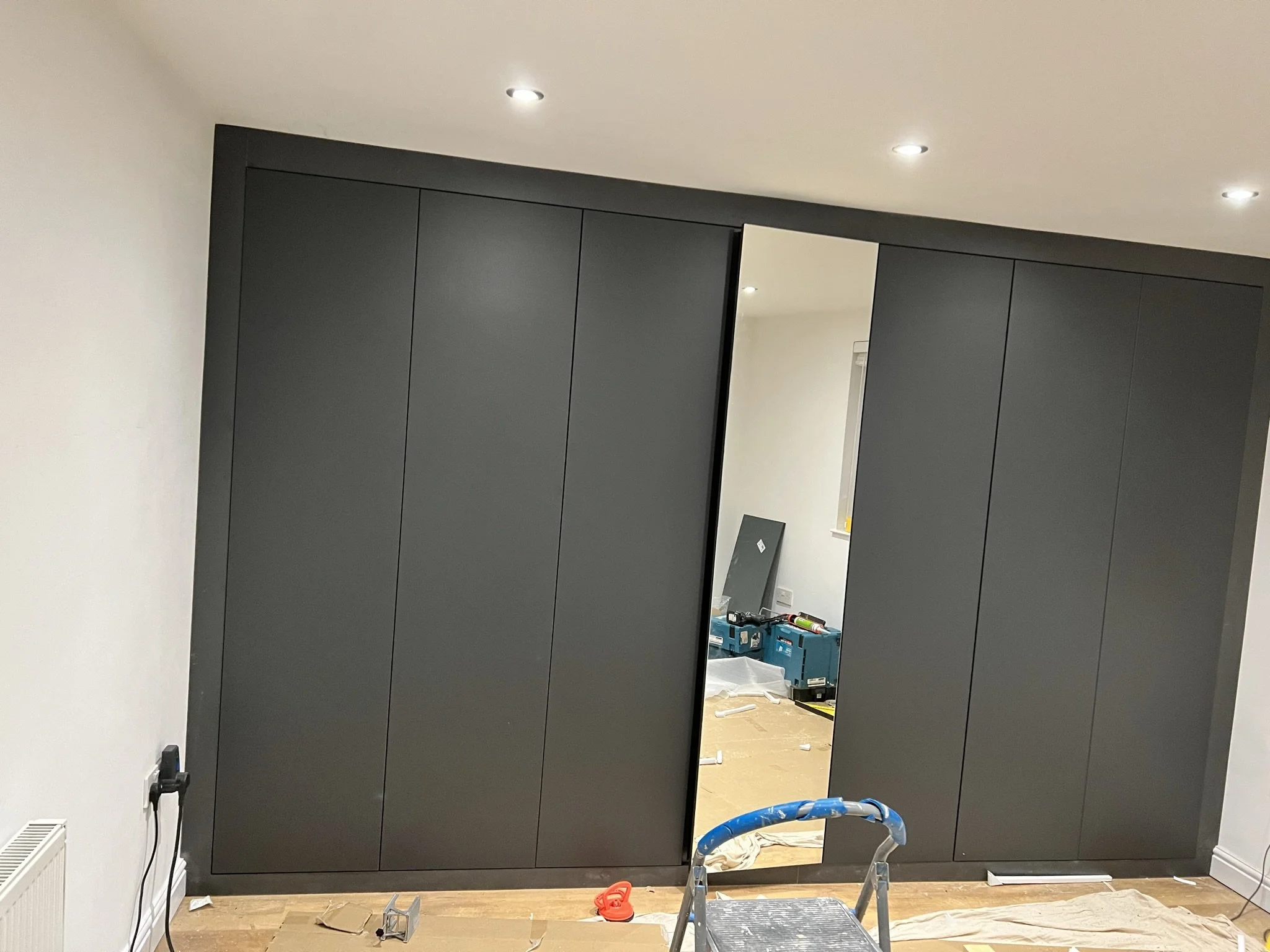Room under construction with a large dark wardrobe, open sliding doors, two-tone flooring, scattered tools, and a stepladder.
