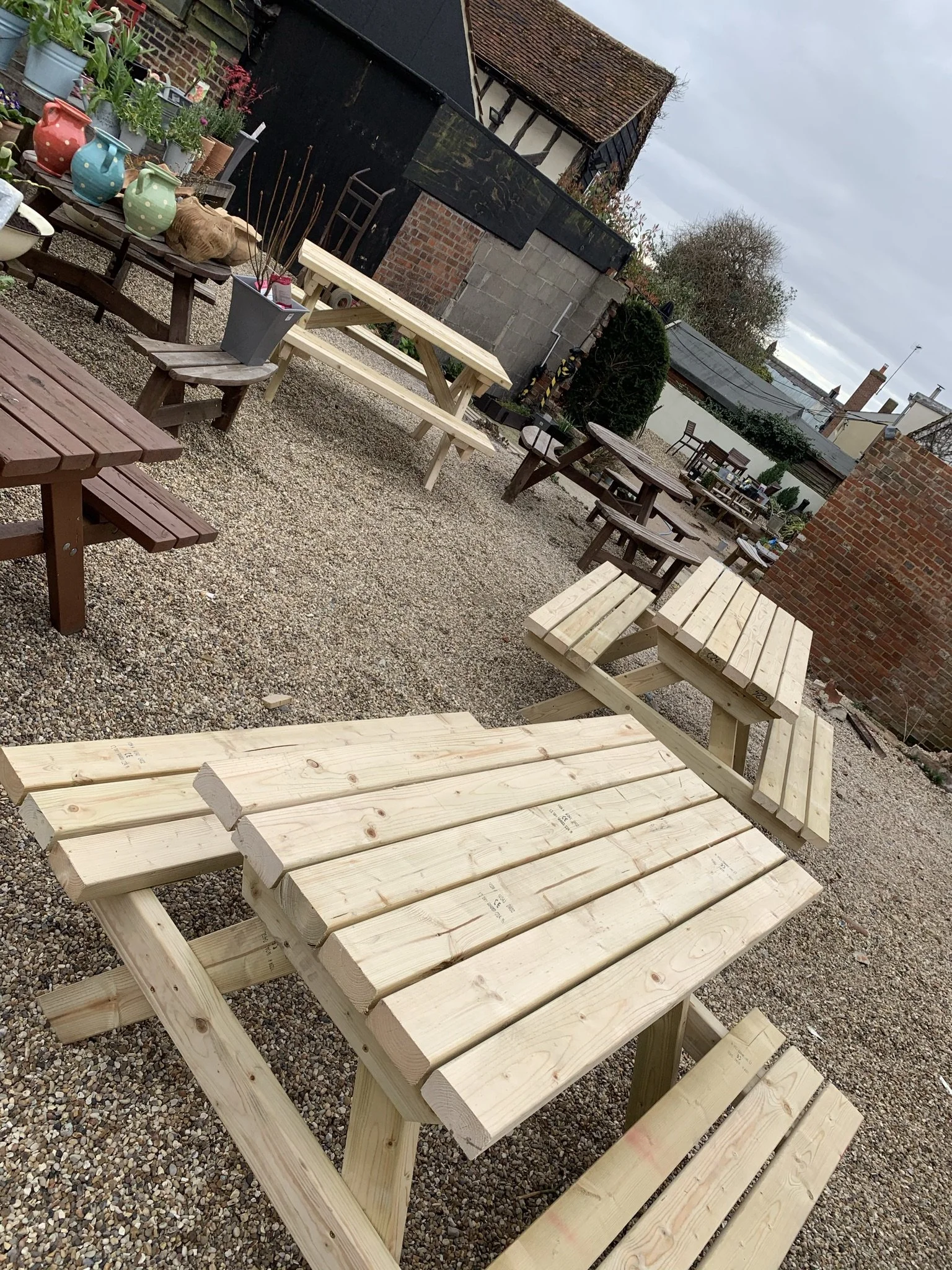 Outdoor picnic tables on gravel surface with potted plants and rustic building in background.