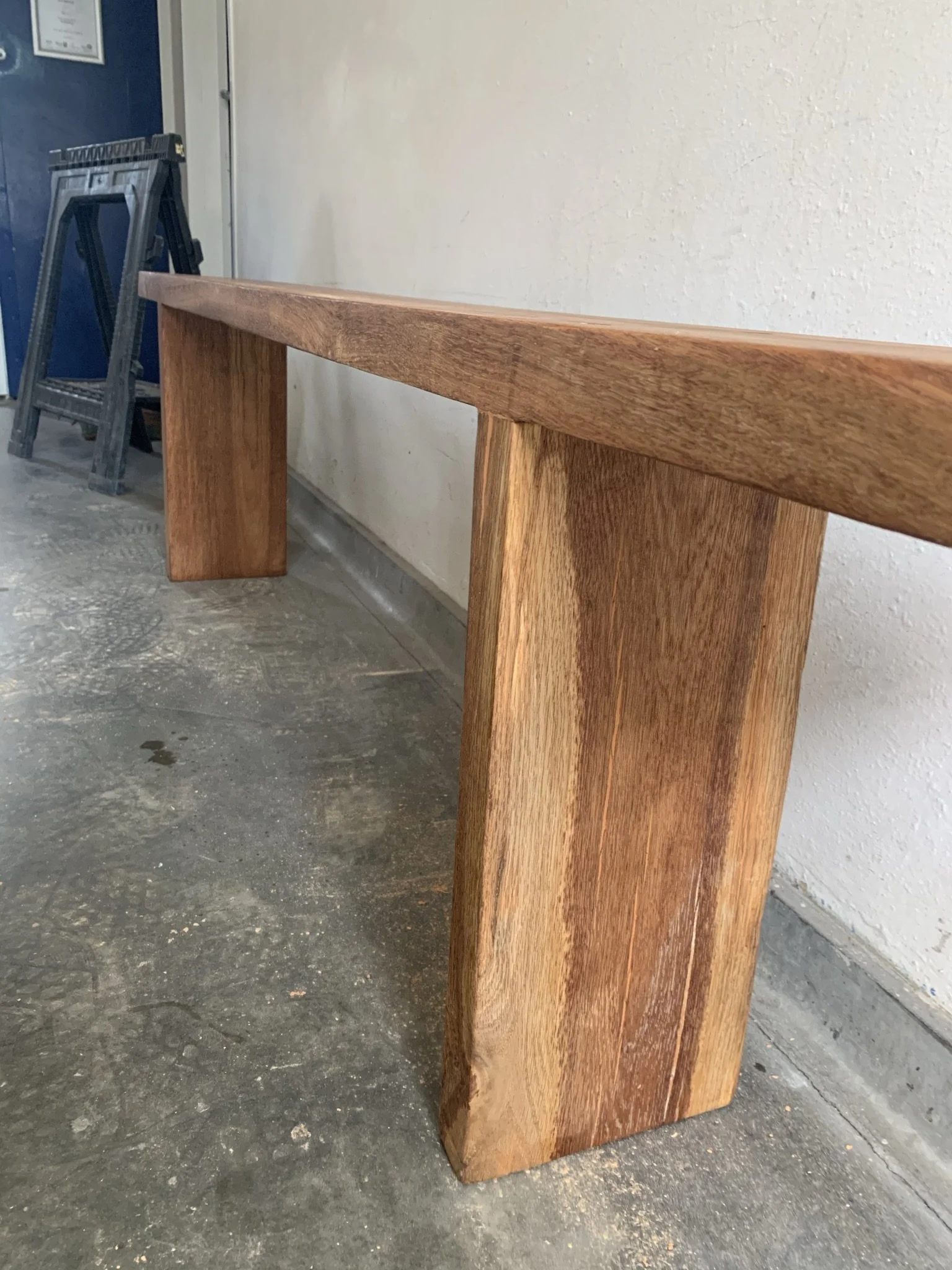 Wooden bench with simple design on concrete floor next to a wall, with a blue door and a folding saw horse in the background.