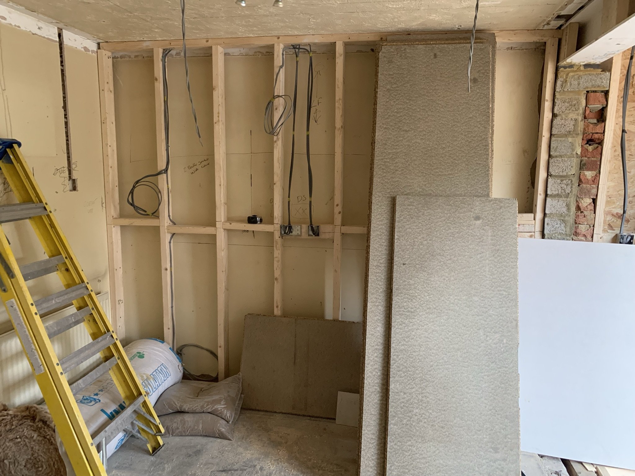 Room under construction with exposed wooden frame, yellow ladder, wall insulation panels, loose wires, and construction materials on the floor.