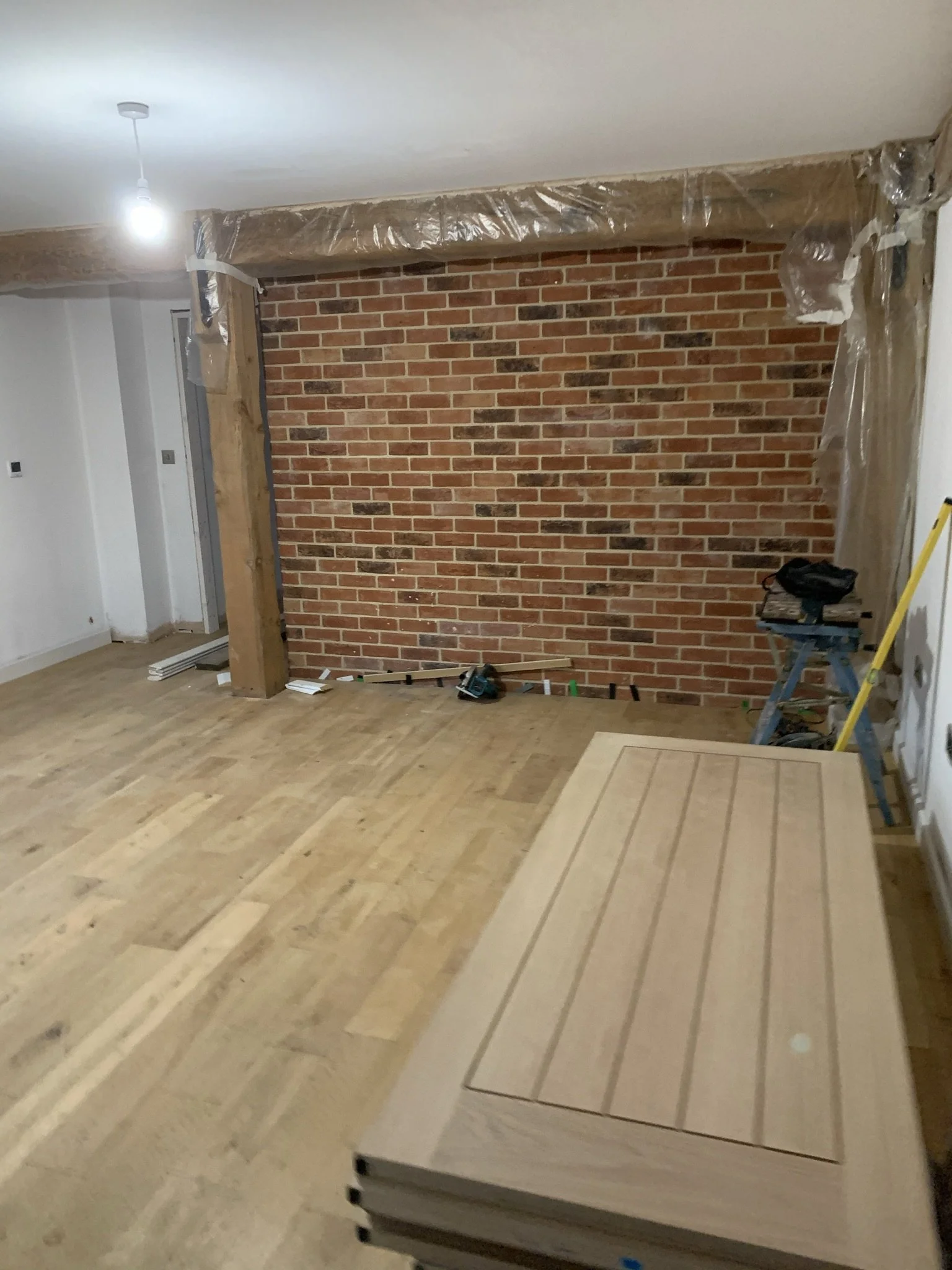 Interior room under renovation with exposed brick wall, wooden floors, stacked doors, and construction materials.