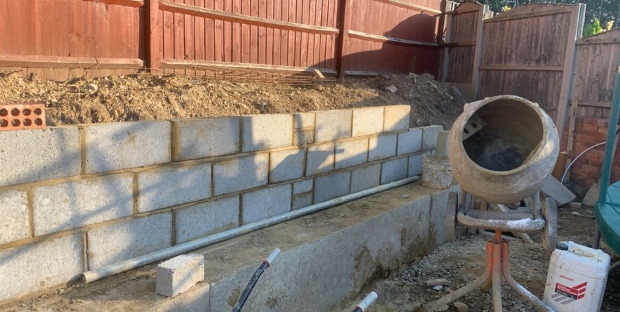 Construction site featuring a partially built concrete block wall with a cement mixer and various construction materials nearby.