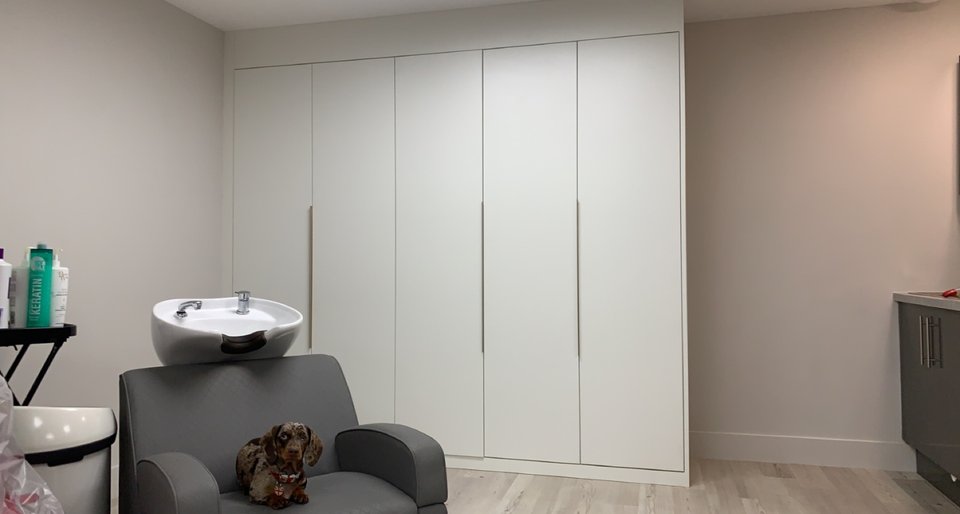 Small dog sitting on a gray salon chair with a hair-washing sink nearby, large white cabinets, and light wooden flooring in a salon or grooming room.