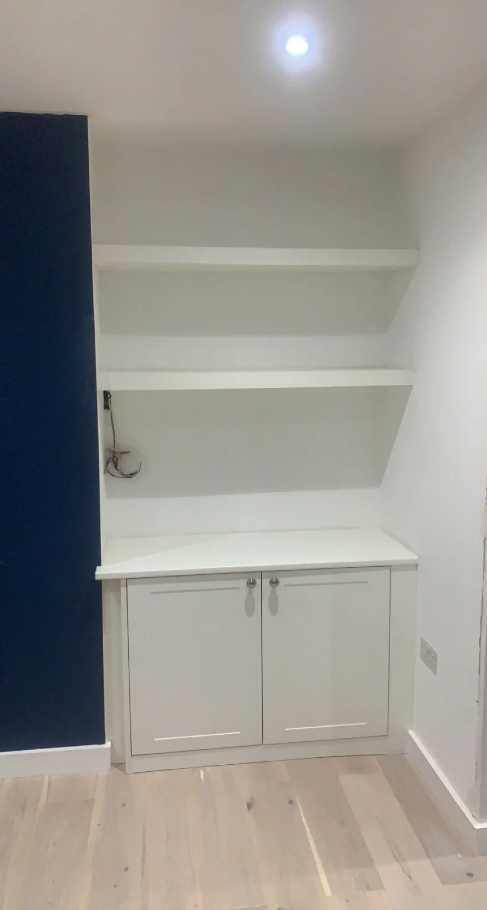 White built-in shelving unit with two open shelves and a cupboard below, next to a navy blue wall and on a wooden floor.