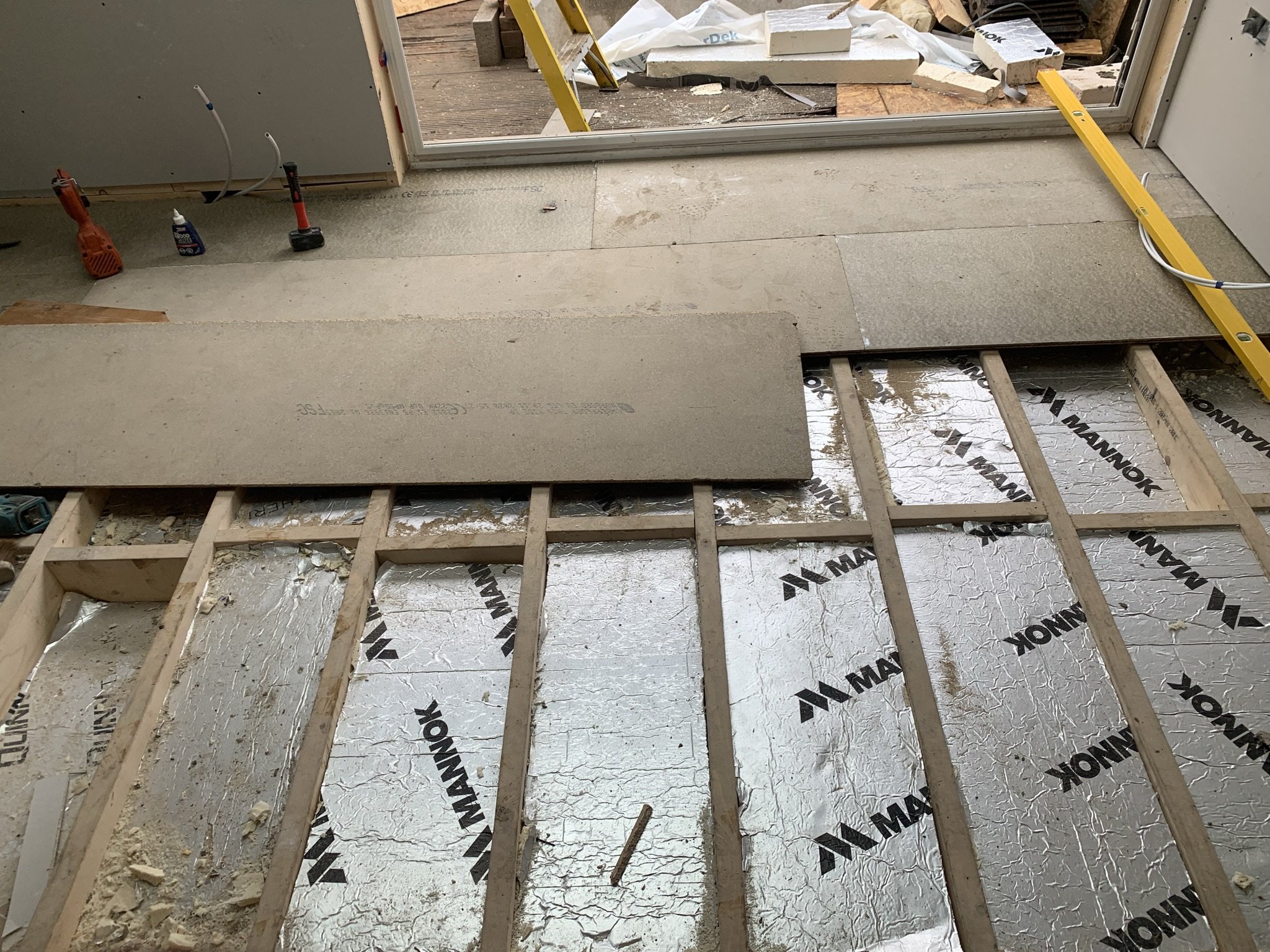 Construction site with exposed floor joists and insulation marked 'Mannok,' partially covered with cement boards, tools scattered around, and a ladder visible in the background.