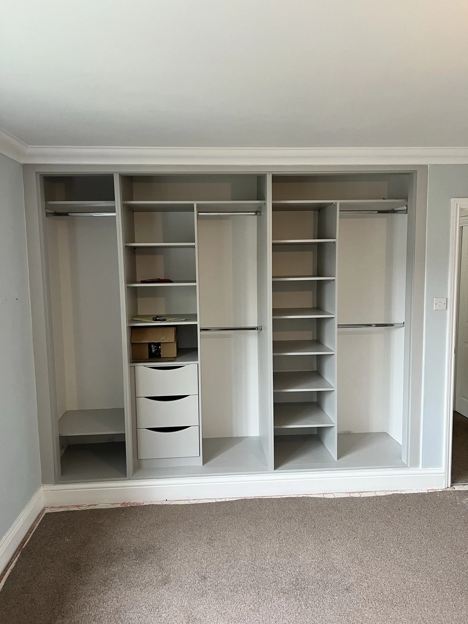 Empty built-in closet with shelves, drawers, and hanging rods in a residential room with gray walls and beige carpet flooring.