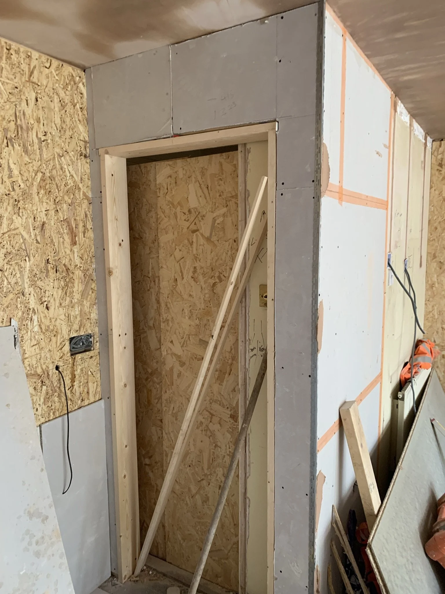 Partially constructed interior wall showing a door frame with wood beams and OSB panels. Various construction materials and tools are visible, including drywall and electrical wires.