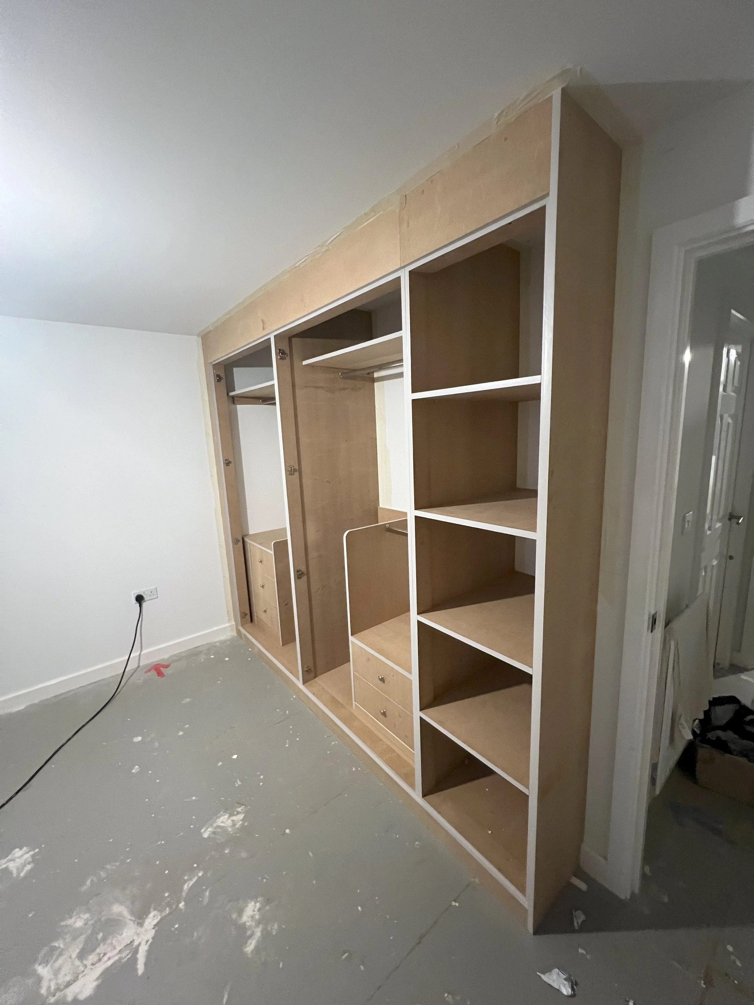 Unfinished built-in wardrobe with open shelves and drawers in a room under renovation, gray concrete floor, and white walls.