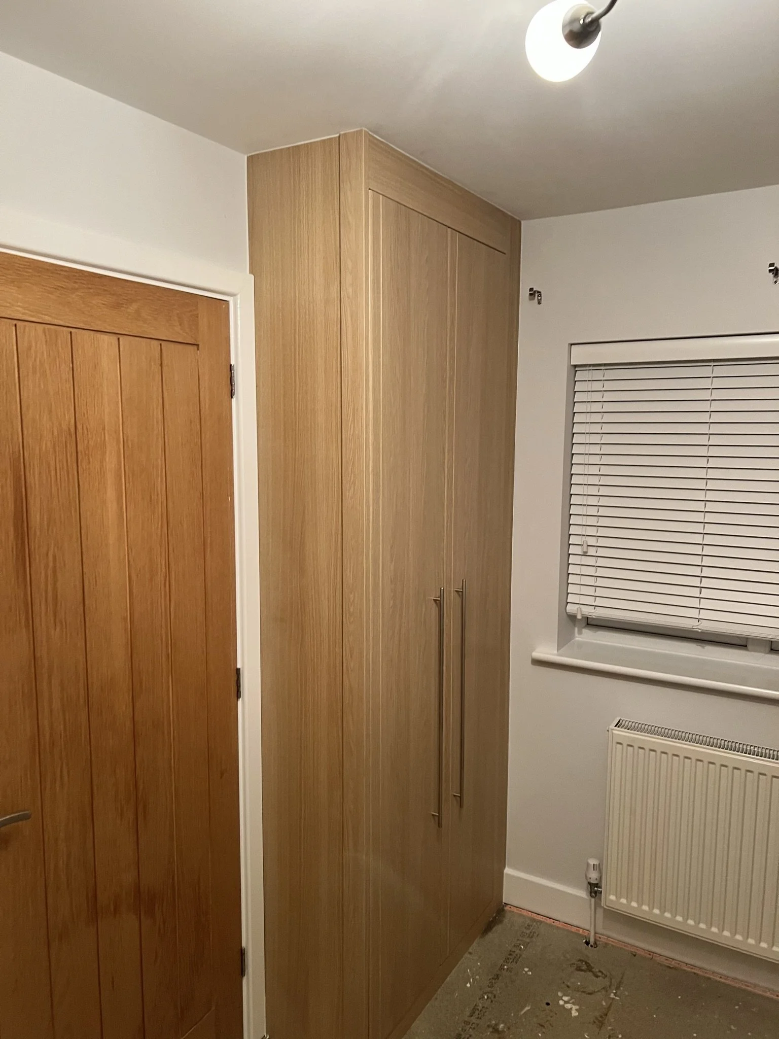 Interior view of a room with a wooden door, a tall wardrobe with two vertical handles, a window with closed blinds, and a radiator below the window. The floor appears unfinished.