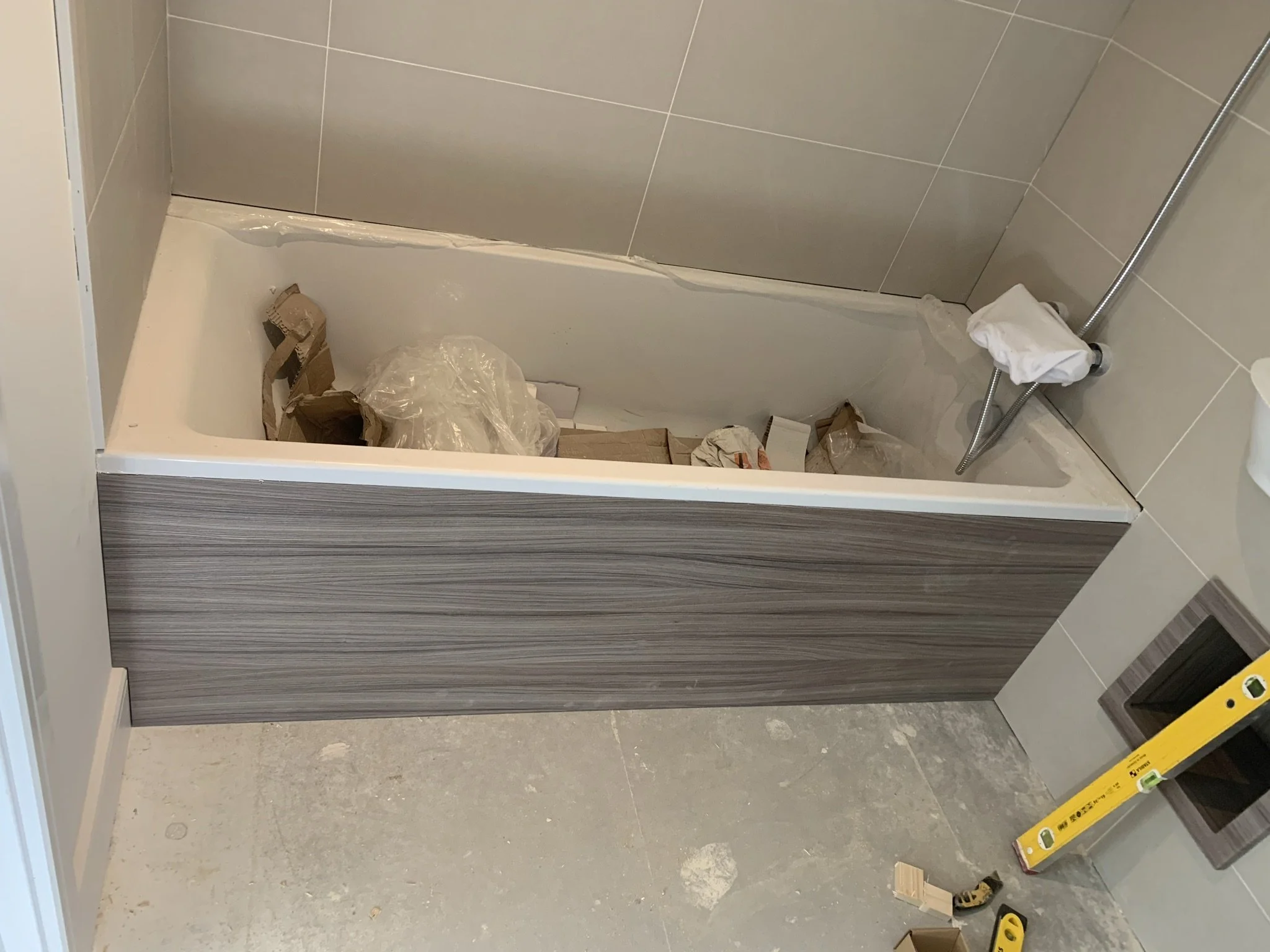 Bathroom under construction with bathtub containing construction materials, tiled walls, and a level tool on the floor.