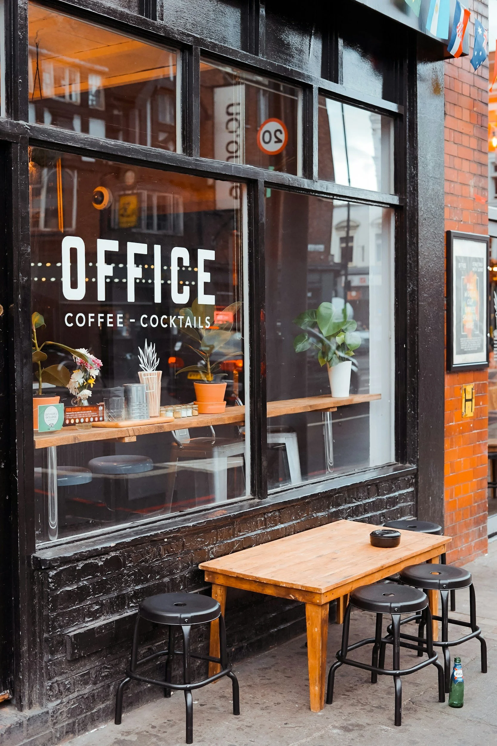 A storefront window with the sign 'OFFICE Coffee Cocktails' on it, decorated with potted plants inside, and an outdoor seating area with a small wooden table, three black stools, and a green bottle on the sidewalk.