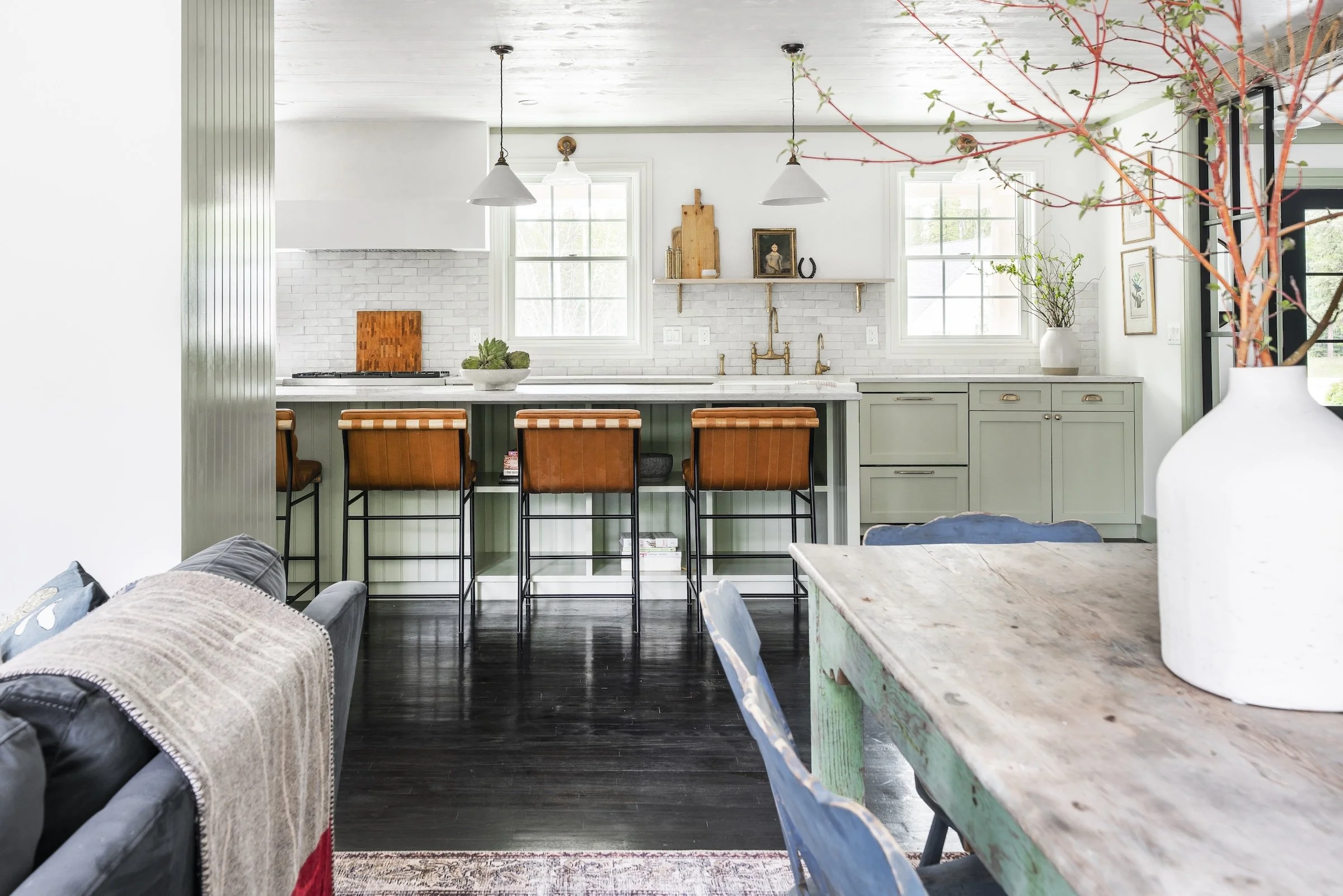 Open-concept kitchen with pale green cabinets, white subway tile backsplash, and a white countertop. Four brown leather barstools are at the kitchen island. Two windows above the sink, pendant lights overhead, and decorative items on a floating shelf. Adjacent to the kitchen, part of a dining table with a white vase and red branches is visible, along with a sofa with a beige throw blanket and a pillow.