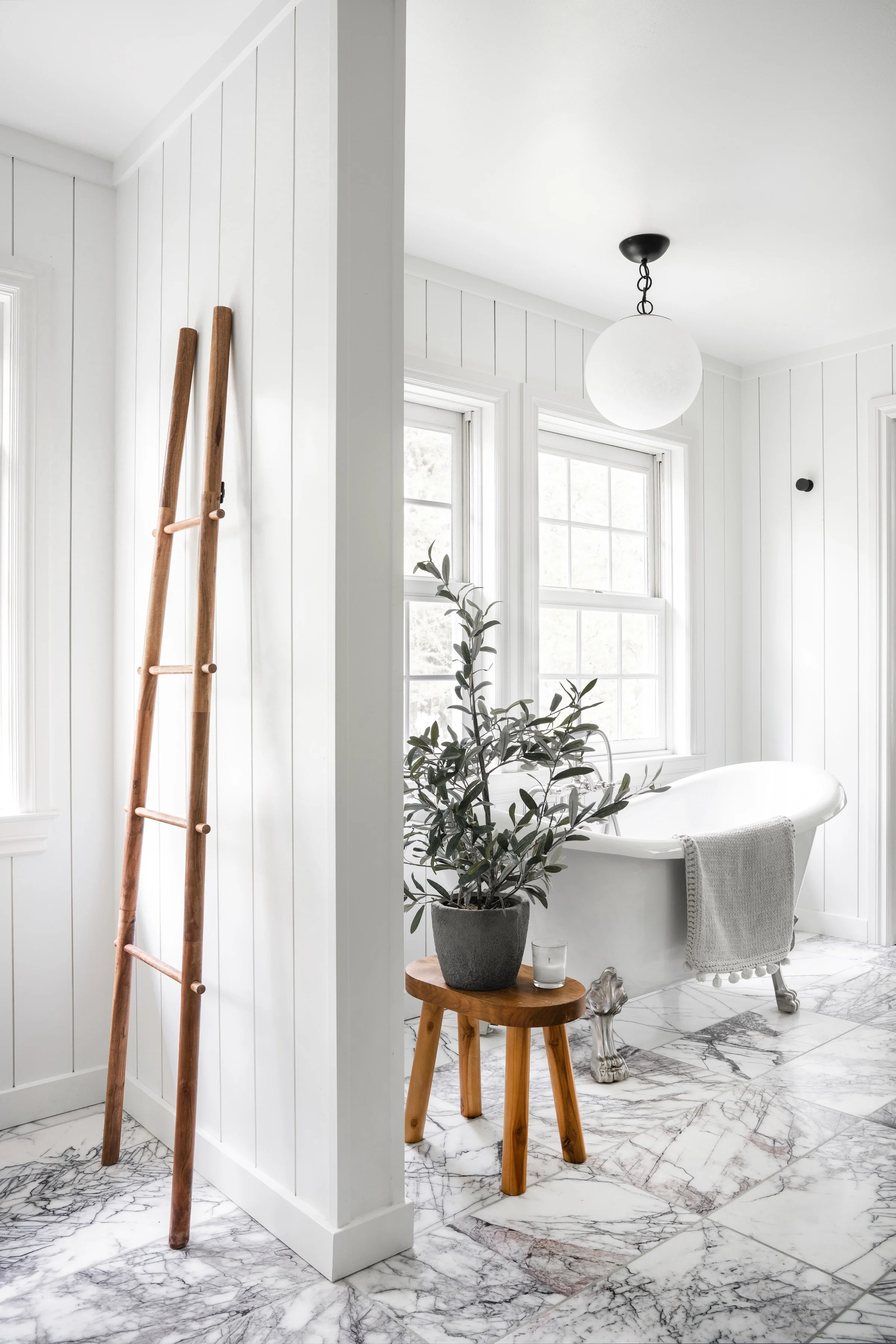 Bright bathroom with clawfoot tub near windows, potted plant, small wooden table, and wooden ladder, all with white walls and marble floors.