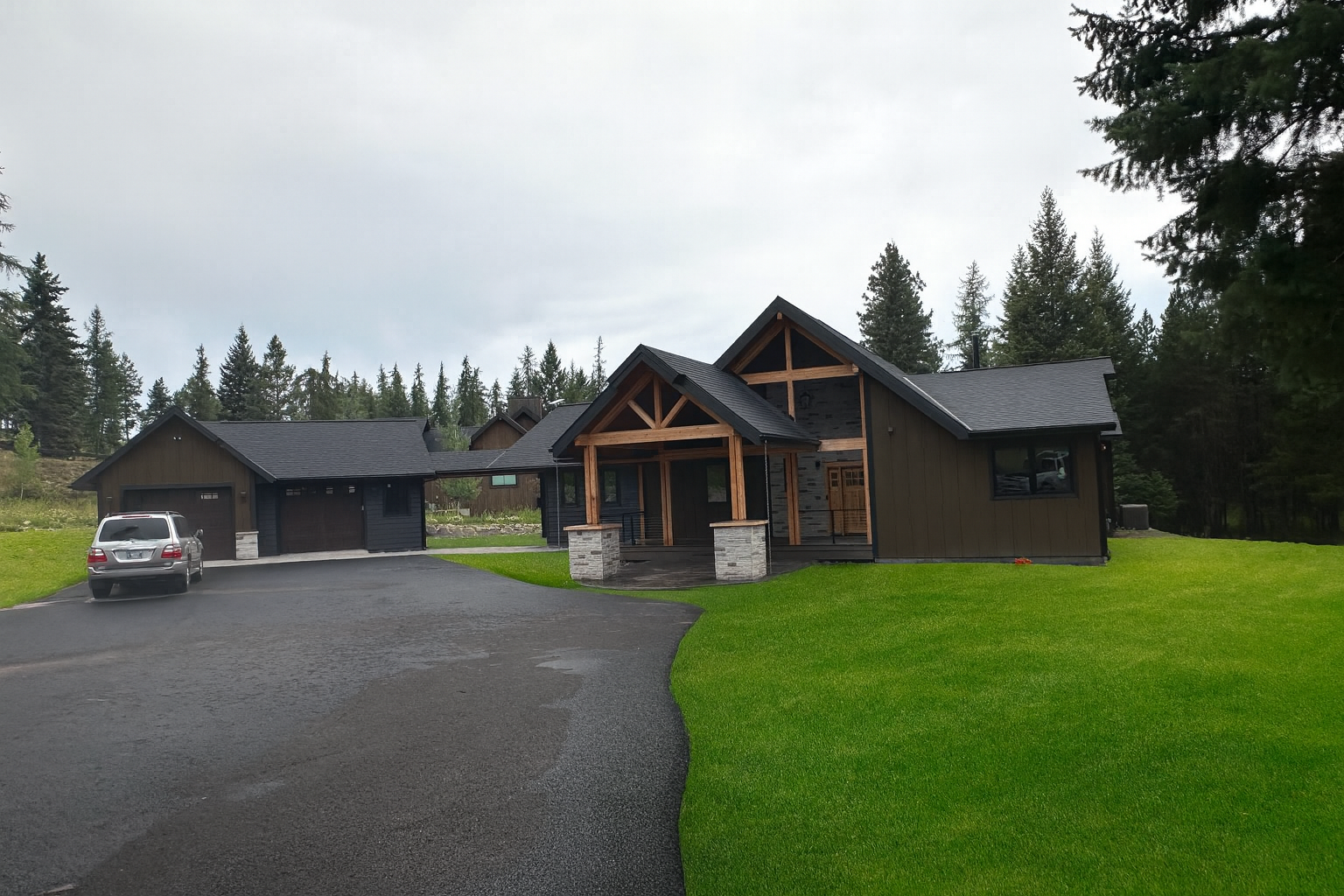A modern house with dark exterior walls, wooden accents, and a stone porch, situated on a lush green lawn with a paved driveway and surrounded by tall trees.
