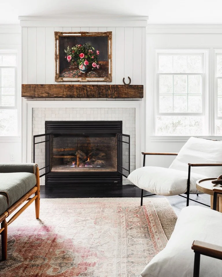 Living room with a fireplace, white walls, a wooden mantel, a framed floral painting above the mantel, a comfy armchair, and large windows providing natural light.