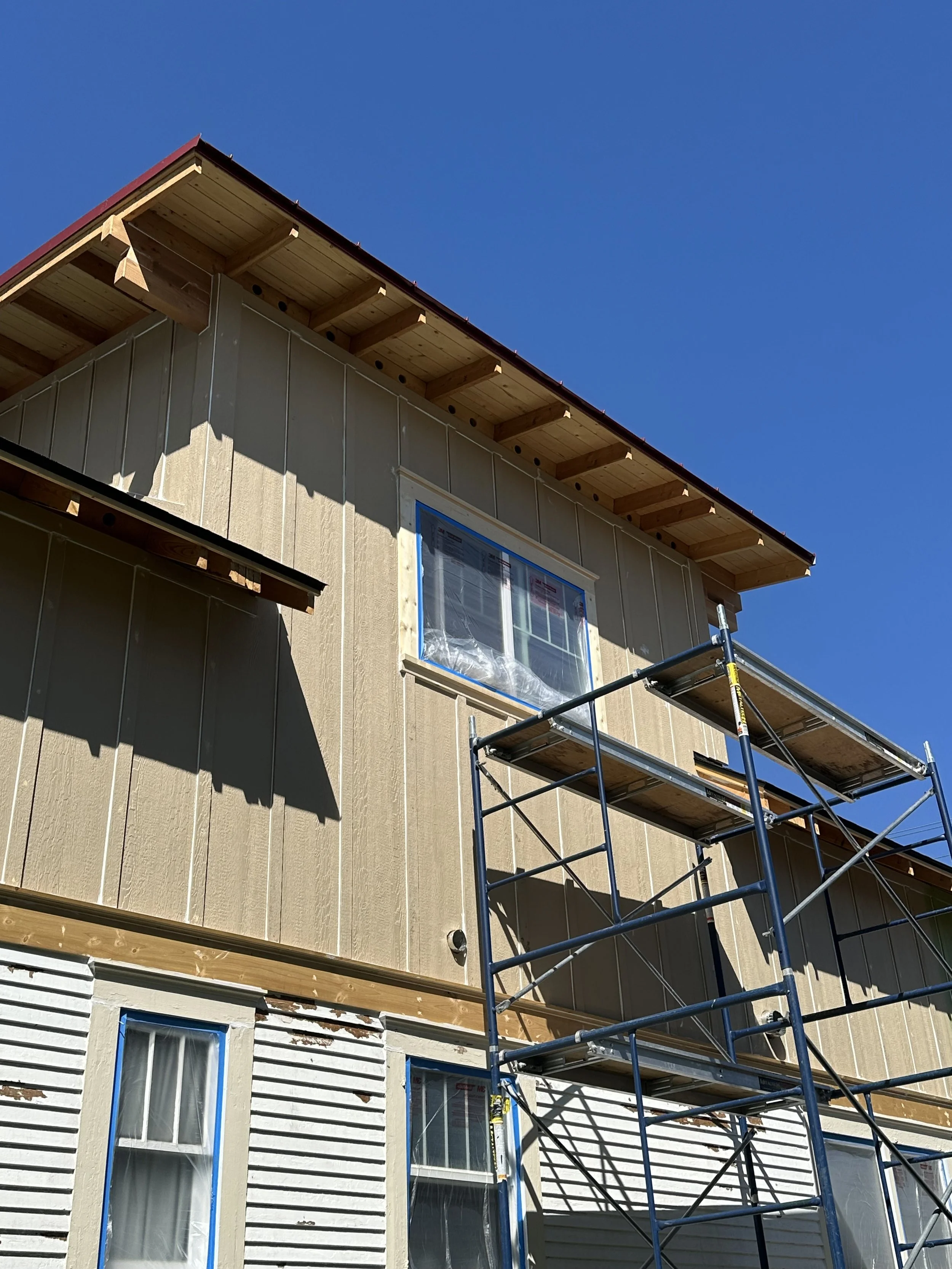 Construction of a house with scaffolding, windows, and wooden siding, under a clear blue sky.