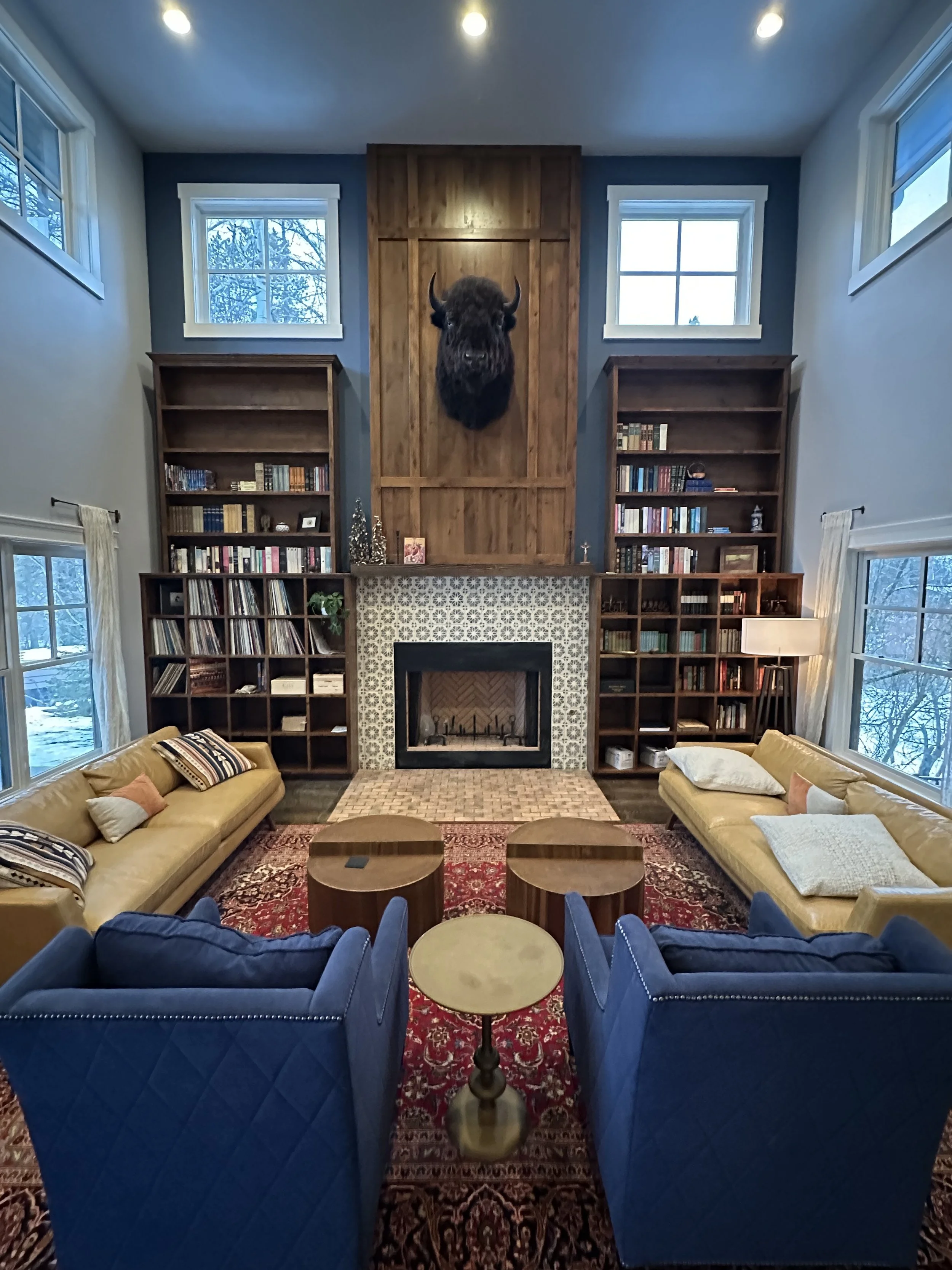 Living room with high ceiling, large windows, a fireplace with black frame and patterned tile surround, built-in bookshelves on either side filled with books and decor, a mounted buffalo head above the fireplace, two beige sofas with pillows, two blue armchairs, a small round table between the armchairs, and a red patterned rug.