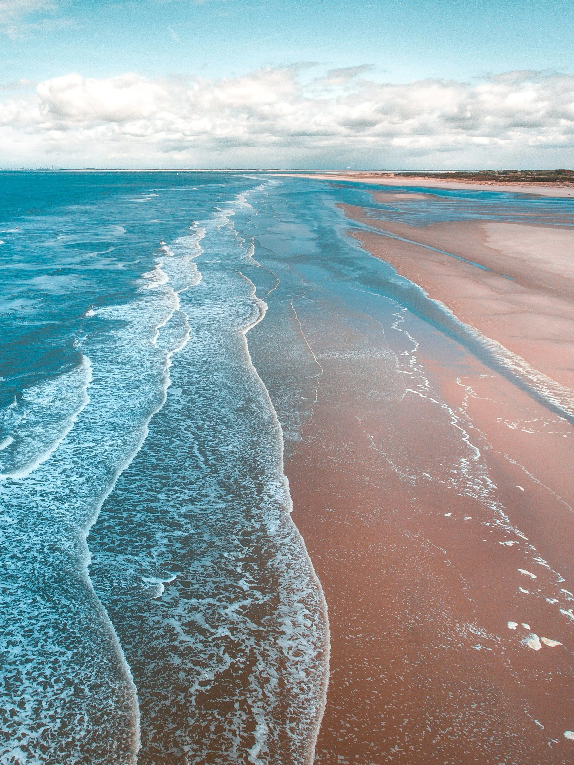 Aerial view of a sandy beach with turquoise ocean waves. The beach is lined with small streams of water flowing towards the sea, under a partly cloudy sky.