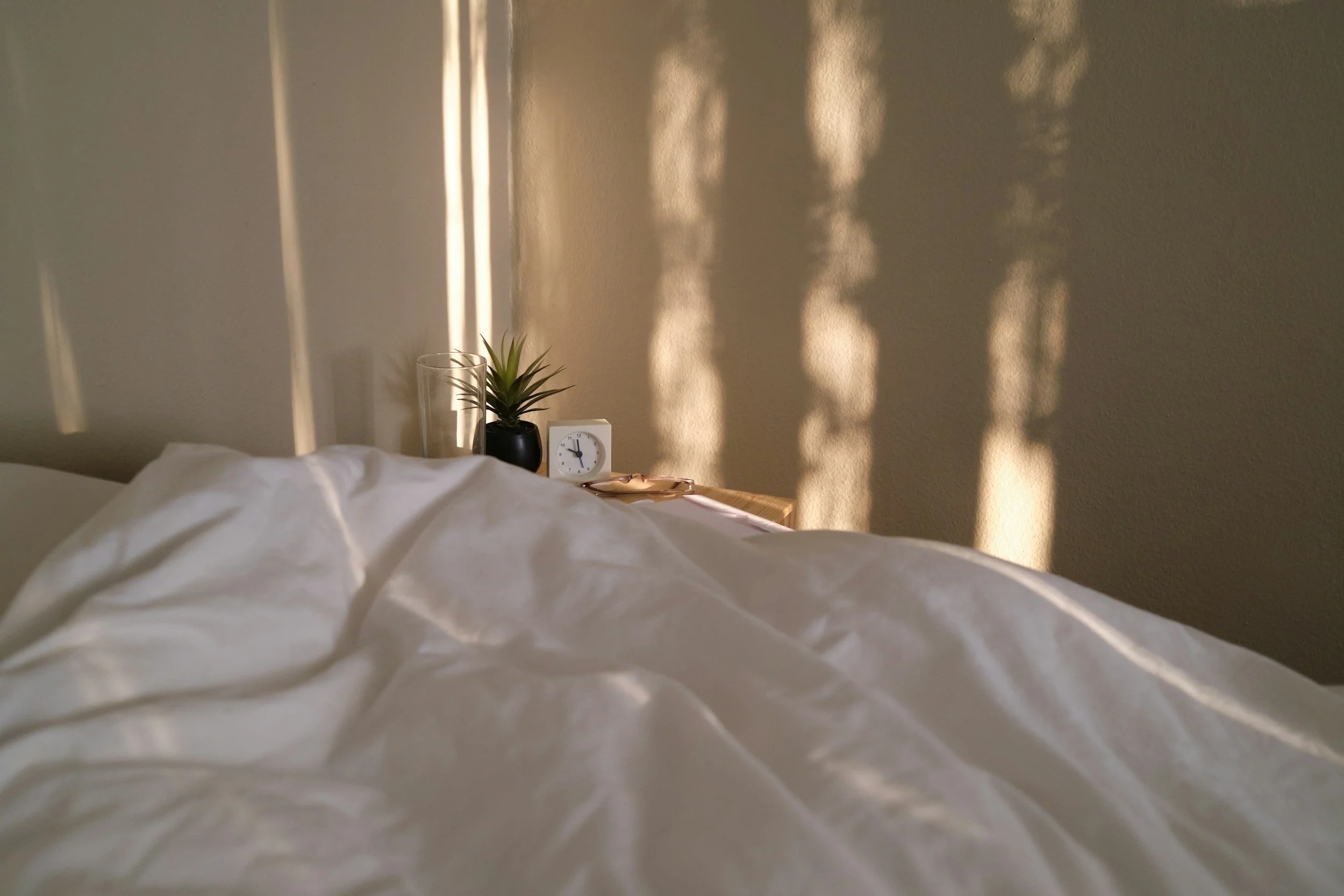 Part of a bed with white sheets, a small wooden bedside table with a white clock, a potted plant in a black pot, and a tall glass. Shadows of leaves are cast on the beige wall in the background, with sunlight coming through blinds.