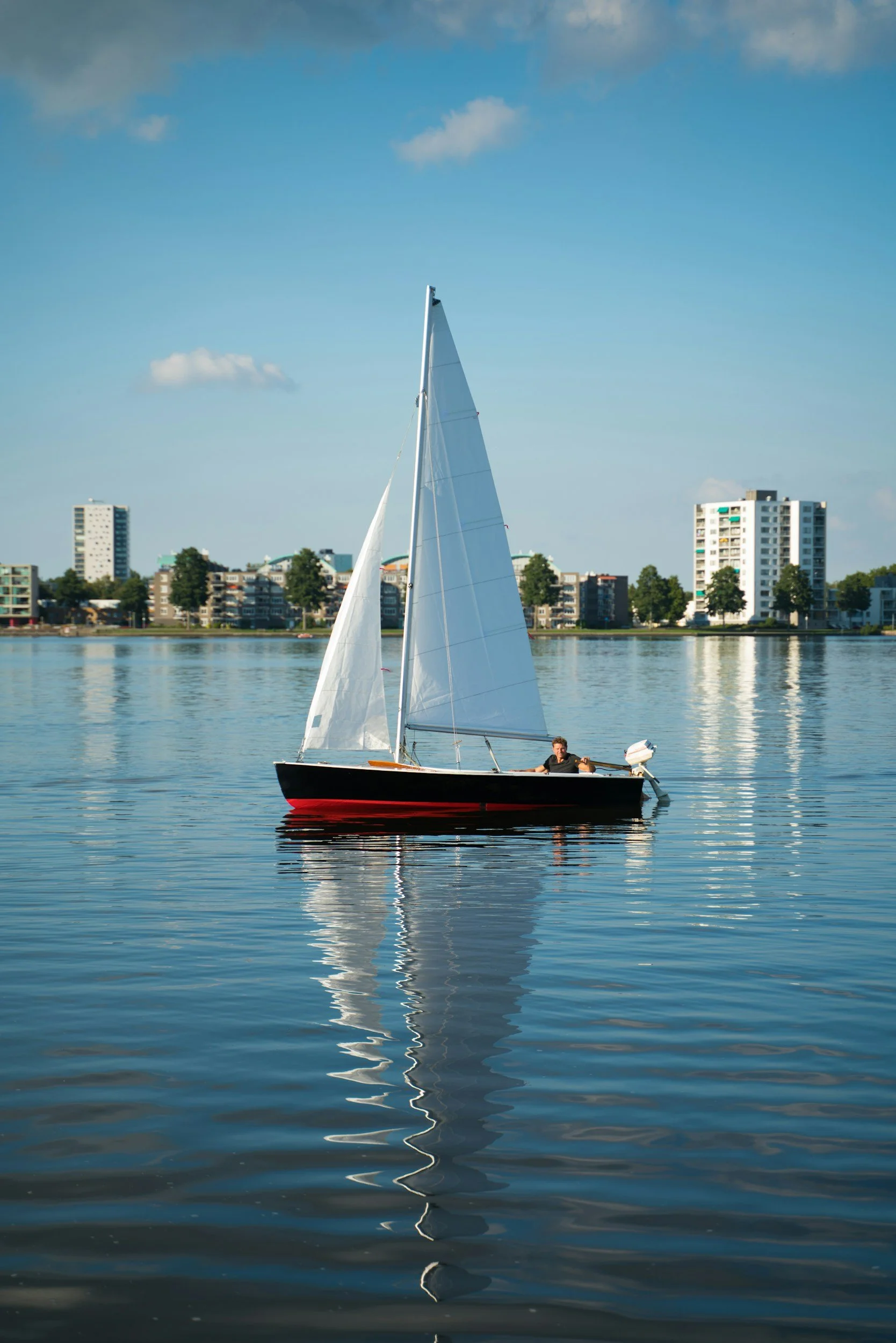 A small sailboat with two white sails floating on calm water, with a city skyline of mid-rise buildings and trees in the background under a blue sky with scattered clouds.