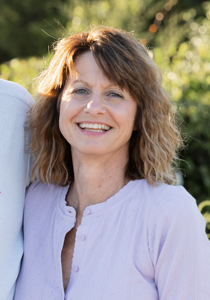 Smiling woman with shoulder-length wavy hair outdoors on a sunny day, wearing a light purple shirt.