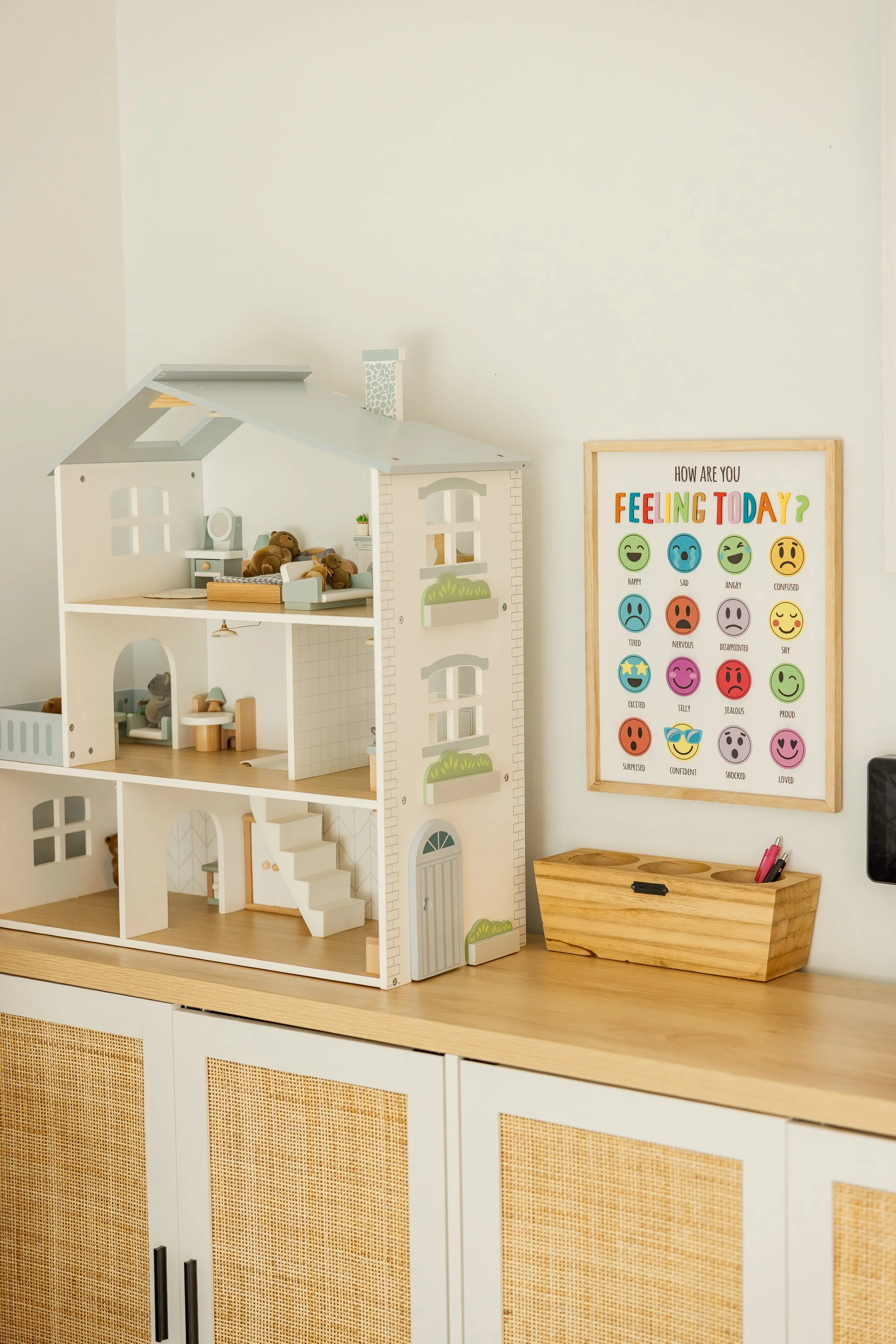 A dollhouse with three floors containing furniture and teddy bears, set on a wooden surface. To the right, a framed chart titled 'How are you feeling today?' shows various colorful emoji expressing different emotions. A wooden box with pens is on the surface beside the chart.