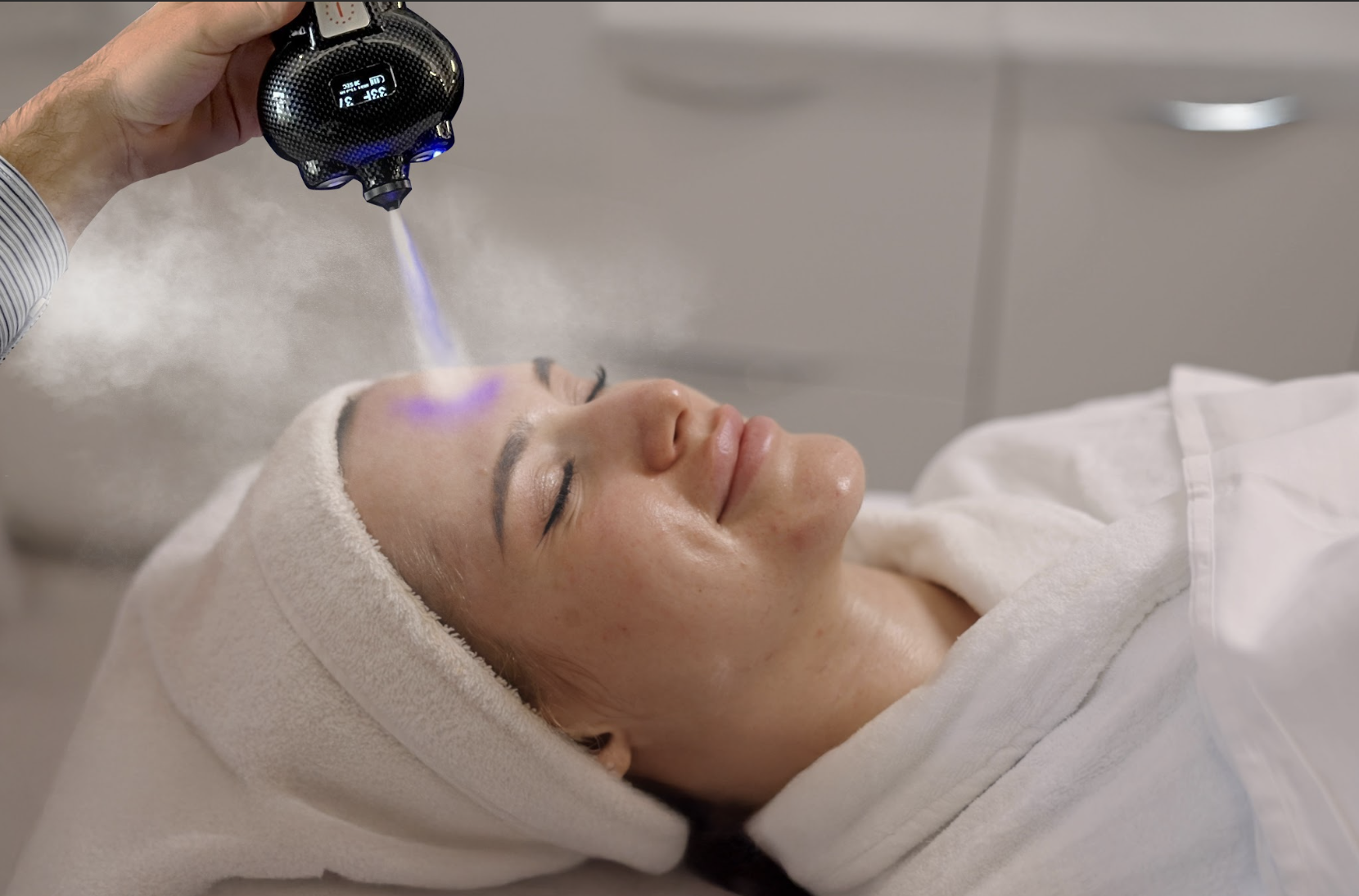 A woman receiving a facial treatment in a spa or clinic, lying on a treatment bed with her eyes closed, wearing a white towel wrapped around her head, while a professional applies a cosmetic device to her forehead.