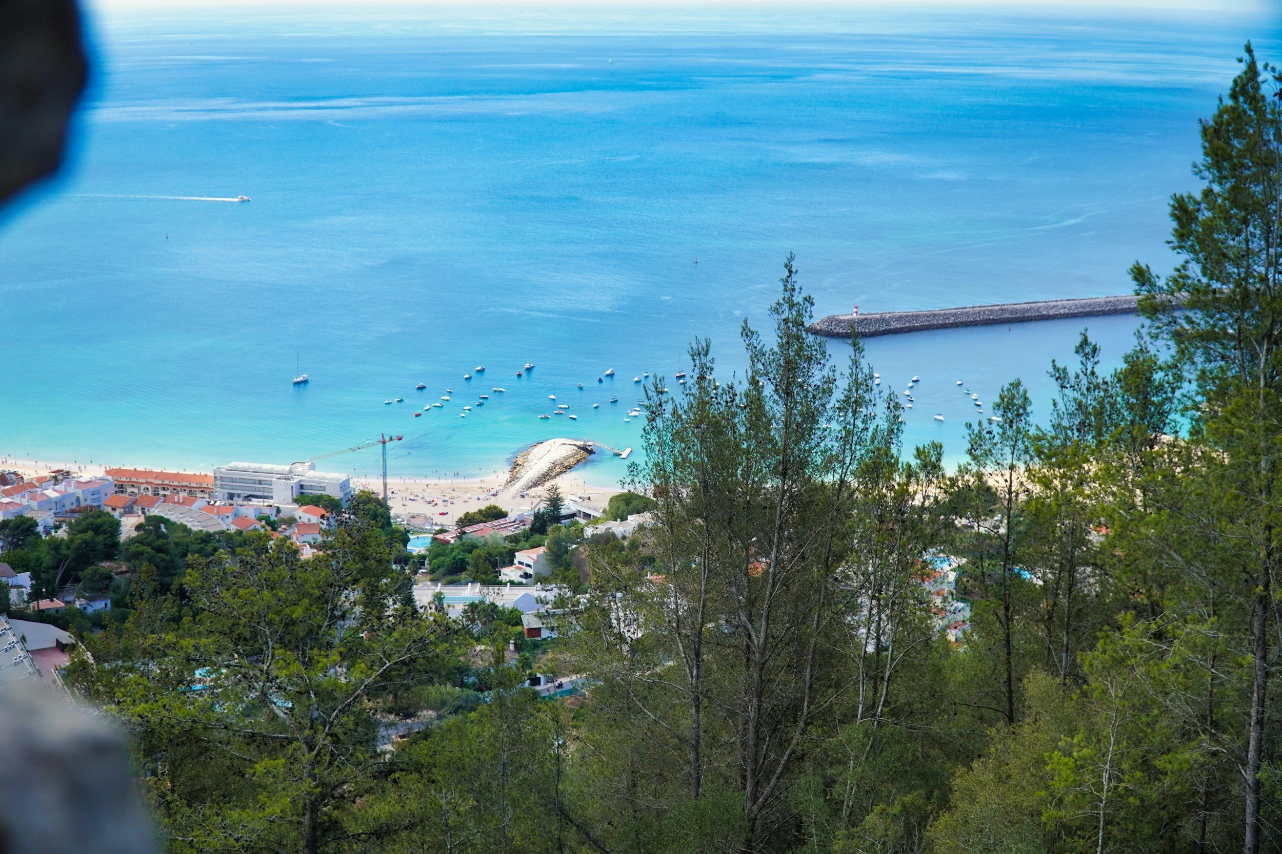 Vista de uma praia com águas azuis claras, barcos ao mar, casas próximas à praia e árvores verdes ao redor, com uma estrutura de contenção no mar.