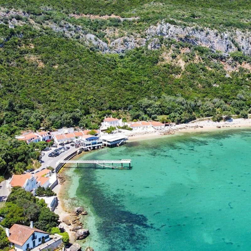 Vista aérea de uma praia com águas transparentes, construída com casas brancas com telhados de barro, cercada por vegetação densa e montanhas ao fundo.