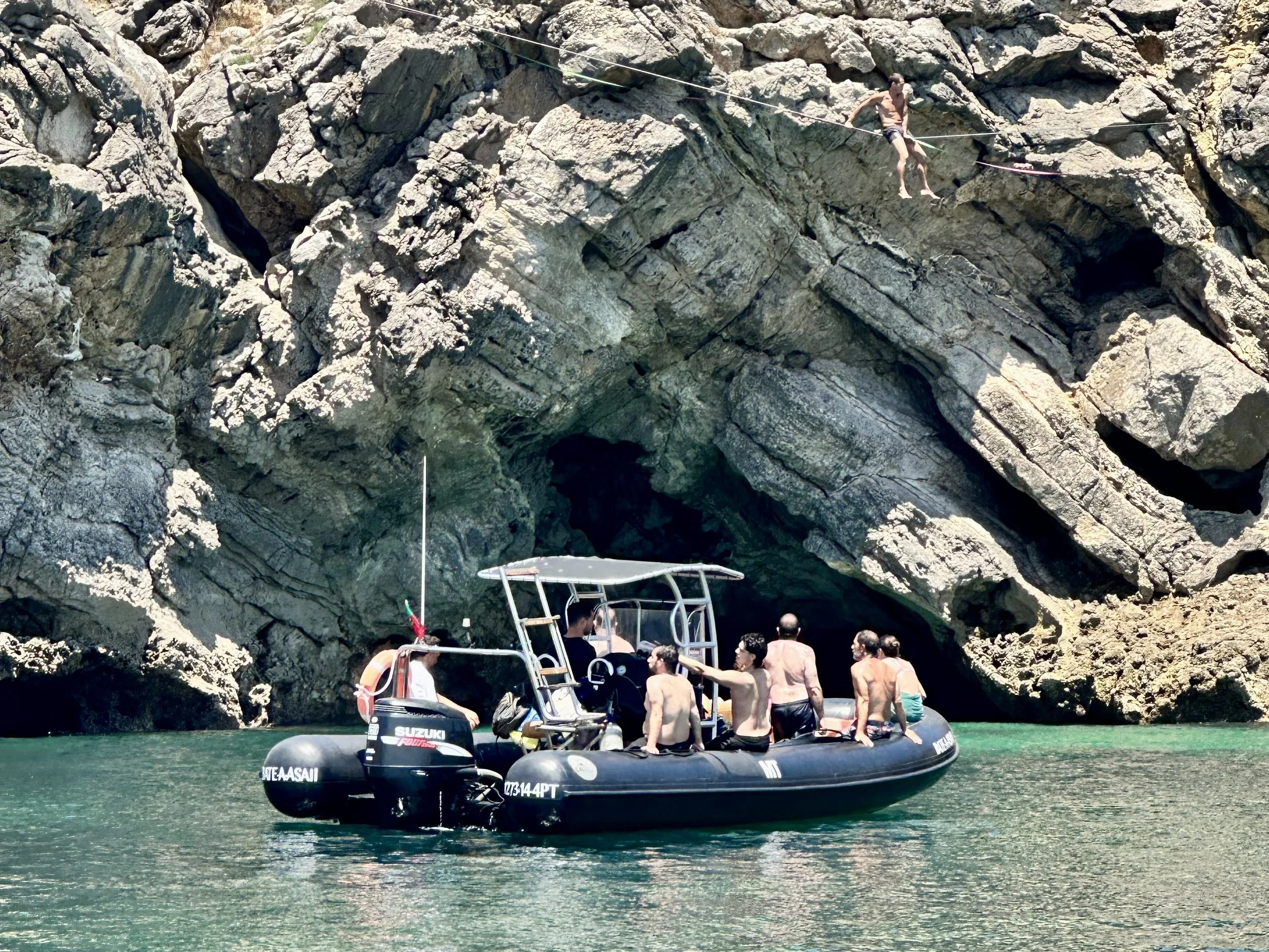 Grupo de pessoas em um barco inflável perto de uma caverna de rochas à beira-mar, com uma pessoa caminhando sobre uma corda presa às rochas.