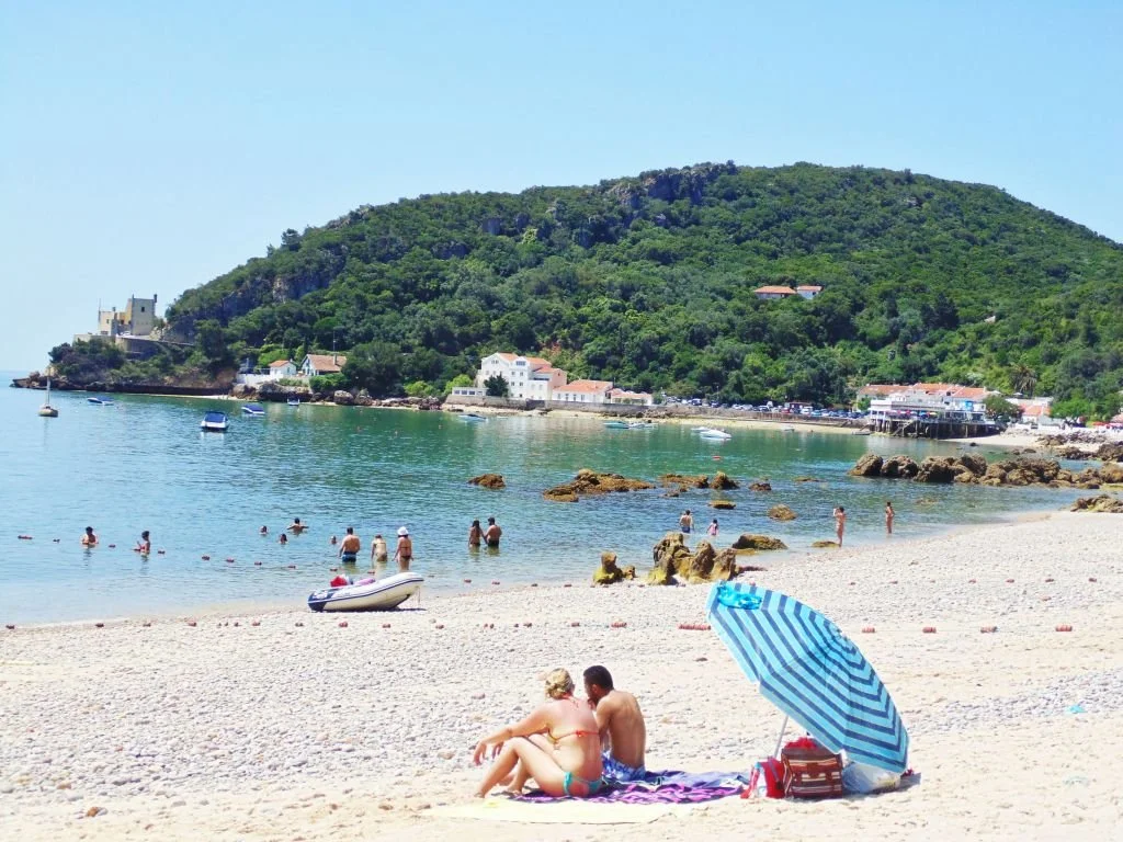 Praia com pessoas na água e na areia, além de um guarda-sol azul e branco na frente, com uma colina verde ao fundo e algumas casas.