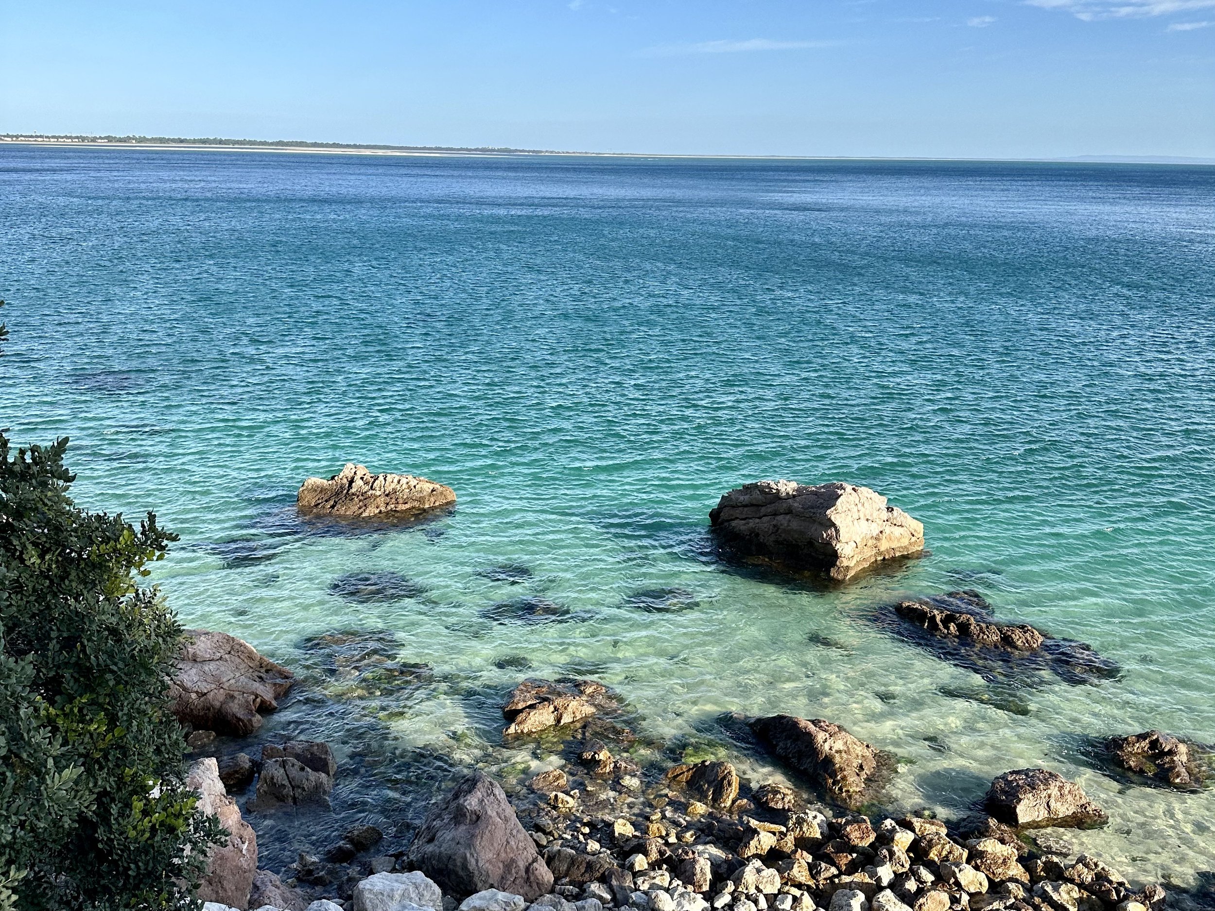 Litoral com água cristalina, rochas na praia e horizonte ao fundo sob céu claro.