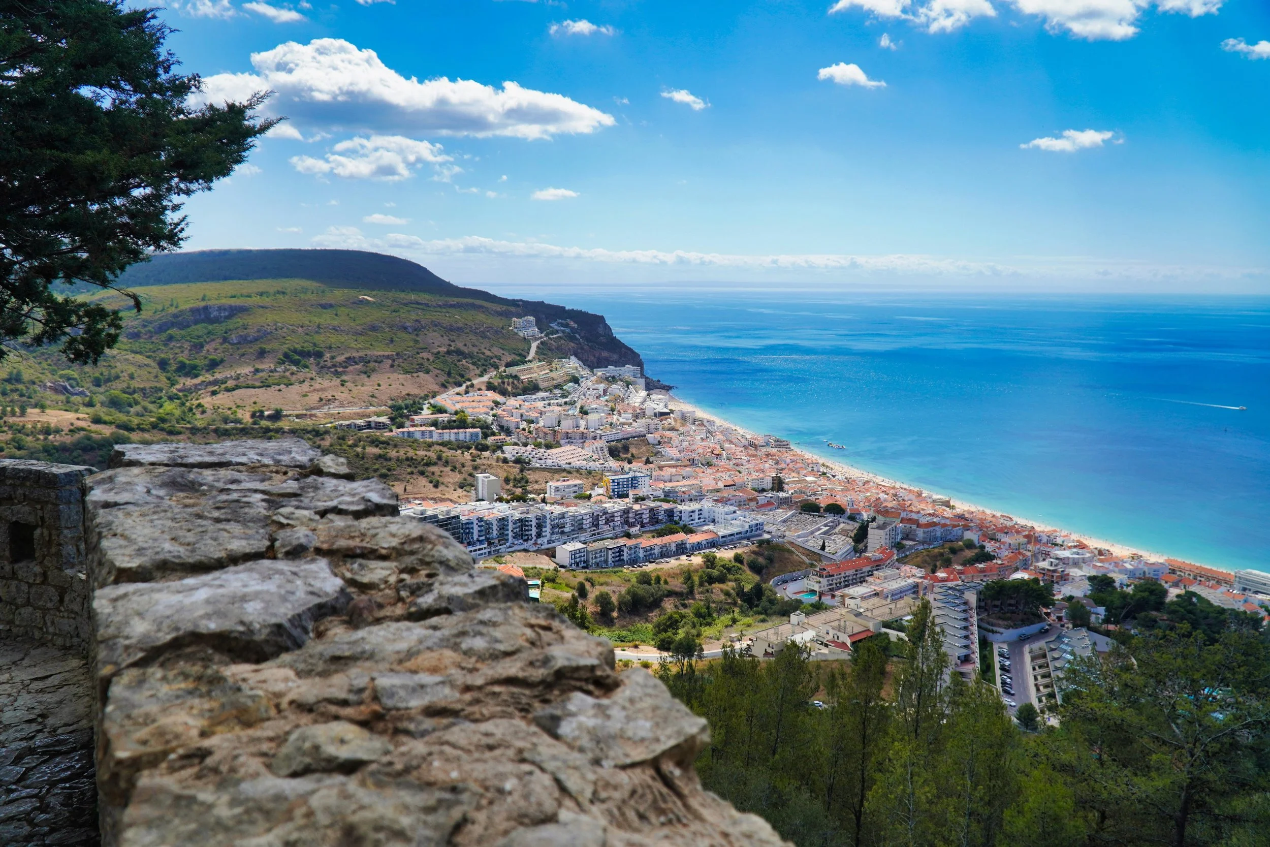 Vista de uma cidade costeira com praias e mar azul, cercada por colinas verdes sob céu azul com nuvens.