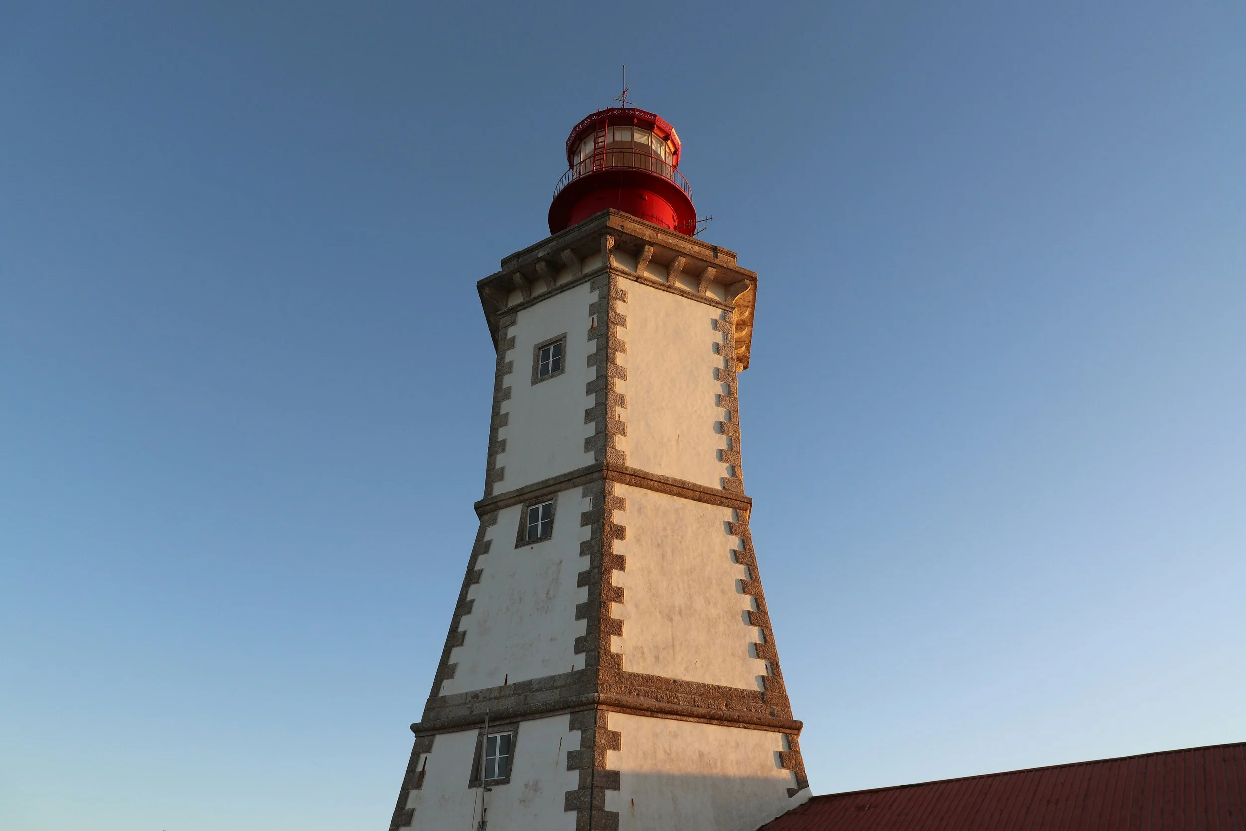Farol branco com detalhes em pedra na esquina, com luz vermelha no topo, contra céu azul.