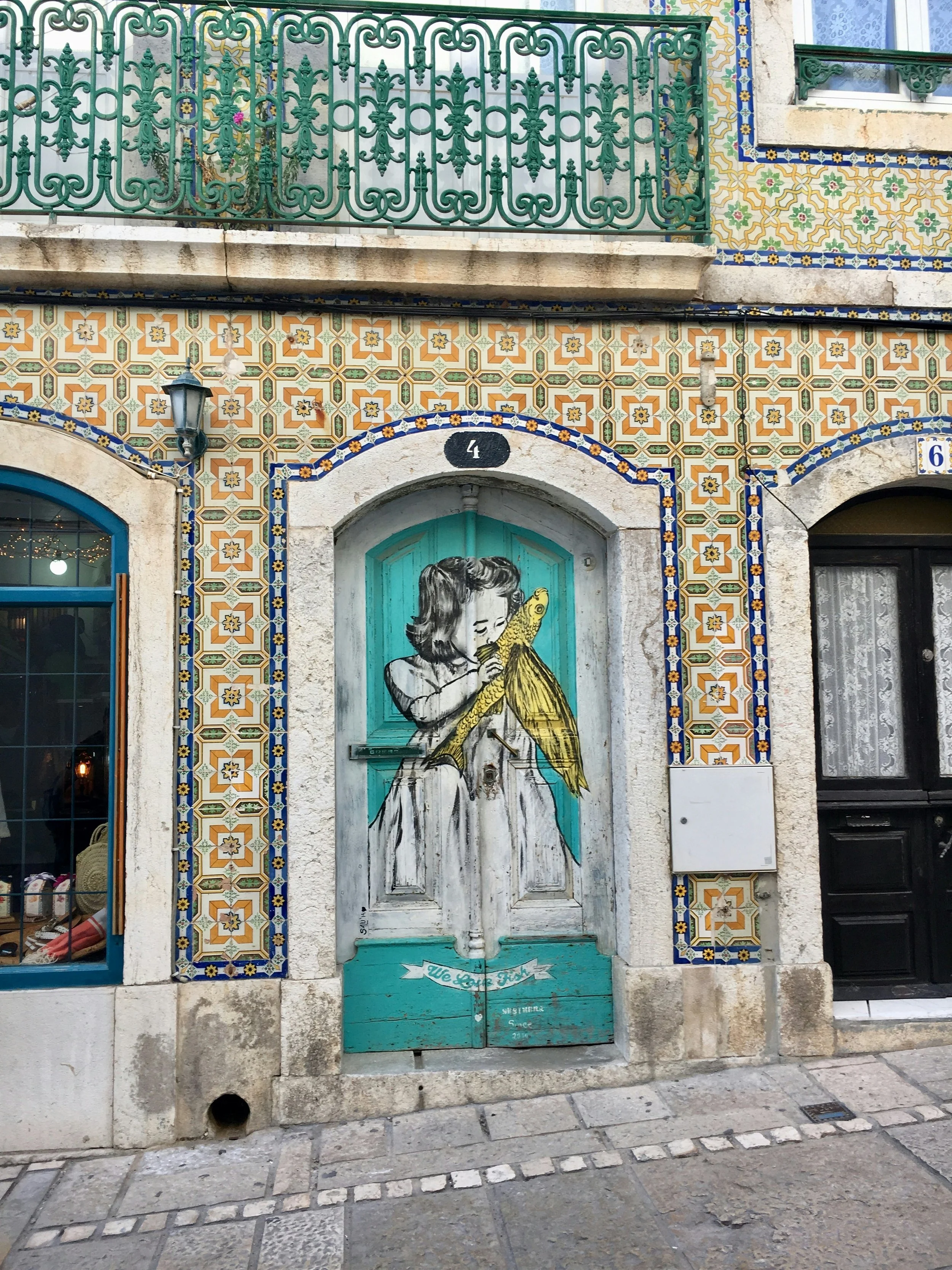 Rua com paredes decoradas com azulejos coloridos. Uma porta de madeira pintada com uma criança desenhada, segurando uma lagosta, com uma faixa que diz "We Love Fish".