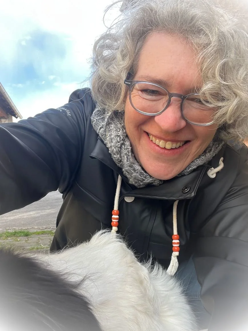 Lächelnde Frau mit Brille, grauen lockigen Haaren, dunkler Jacke und grauem Schal, die ein Tier, vermutlich ein Hund, fotografiert. Der Himmel ist leicht bewölkt.
