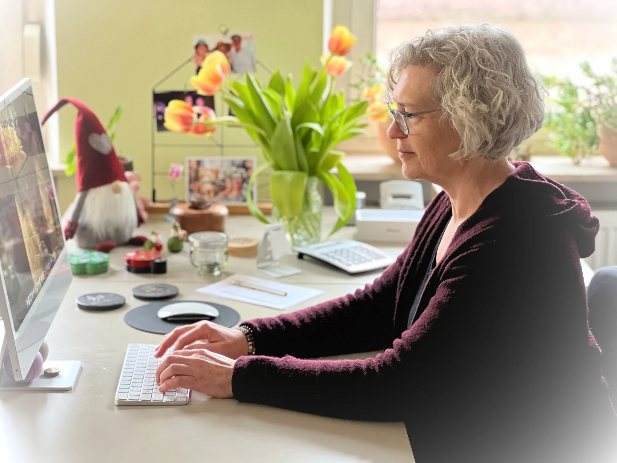Eine ältere Frau mit Locken und Brille arbeitet an einem Computer in einem hellen Raum, der mit bunten Tulpen und einigen Dekorationsgegenständen dekoriert ist.