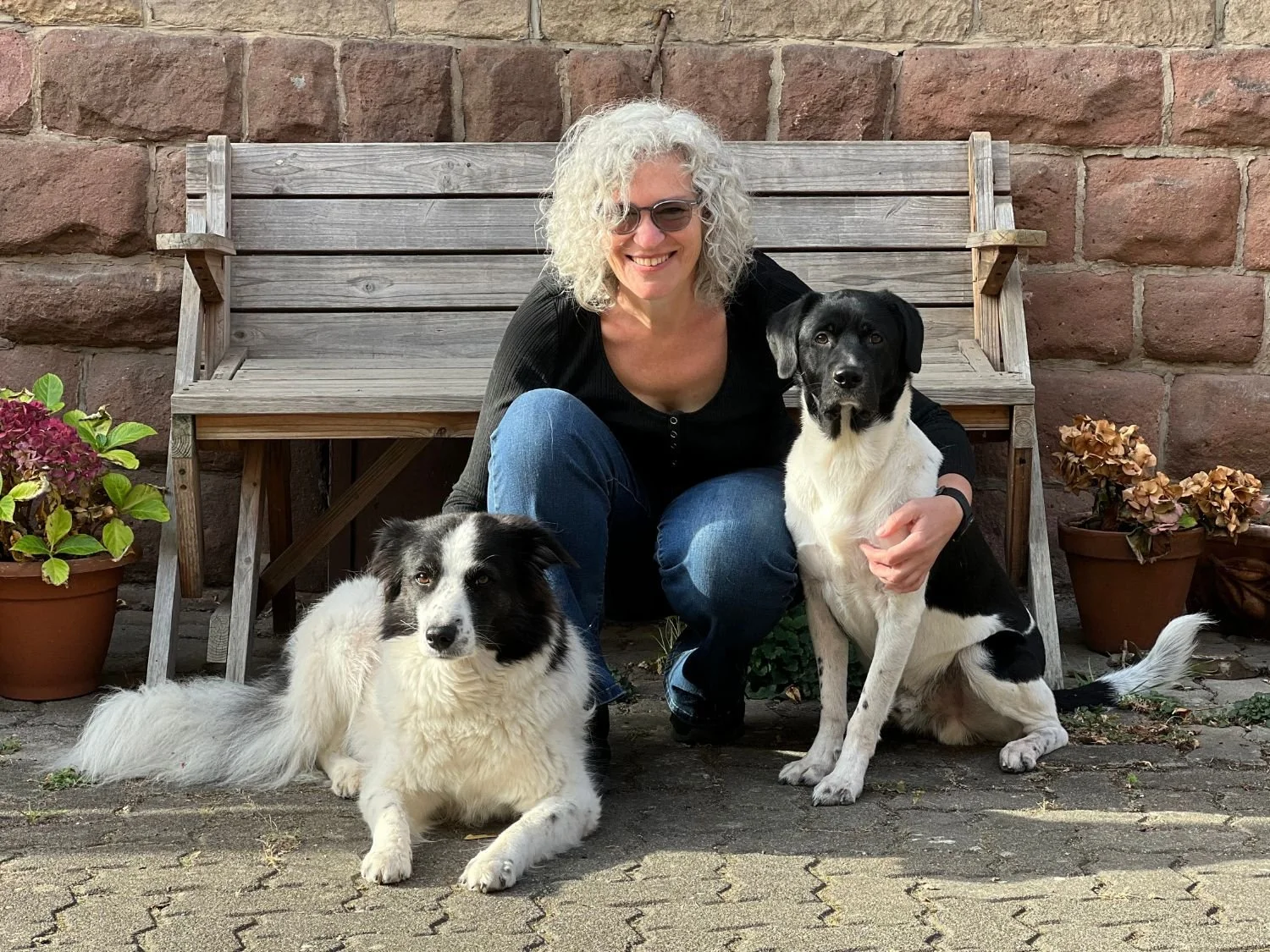 Eine Frau mit lockigem, grauem Haar sitzt auf dem Boden vor einem Holzbänkle, umgeben von zwei Hunden, vor einer Ziegelsteinmauer. Sie lächelt, trägt eine schwarze Oberteil und blaue Jeans. Es sind zwei Blumentöpfe mit Blumen an den Seiten.
