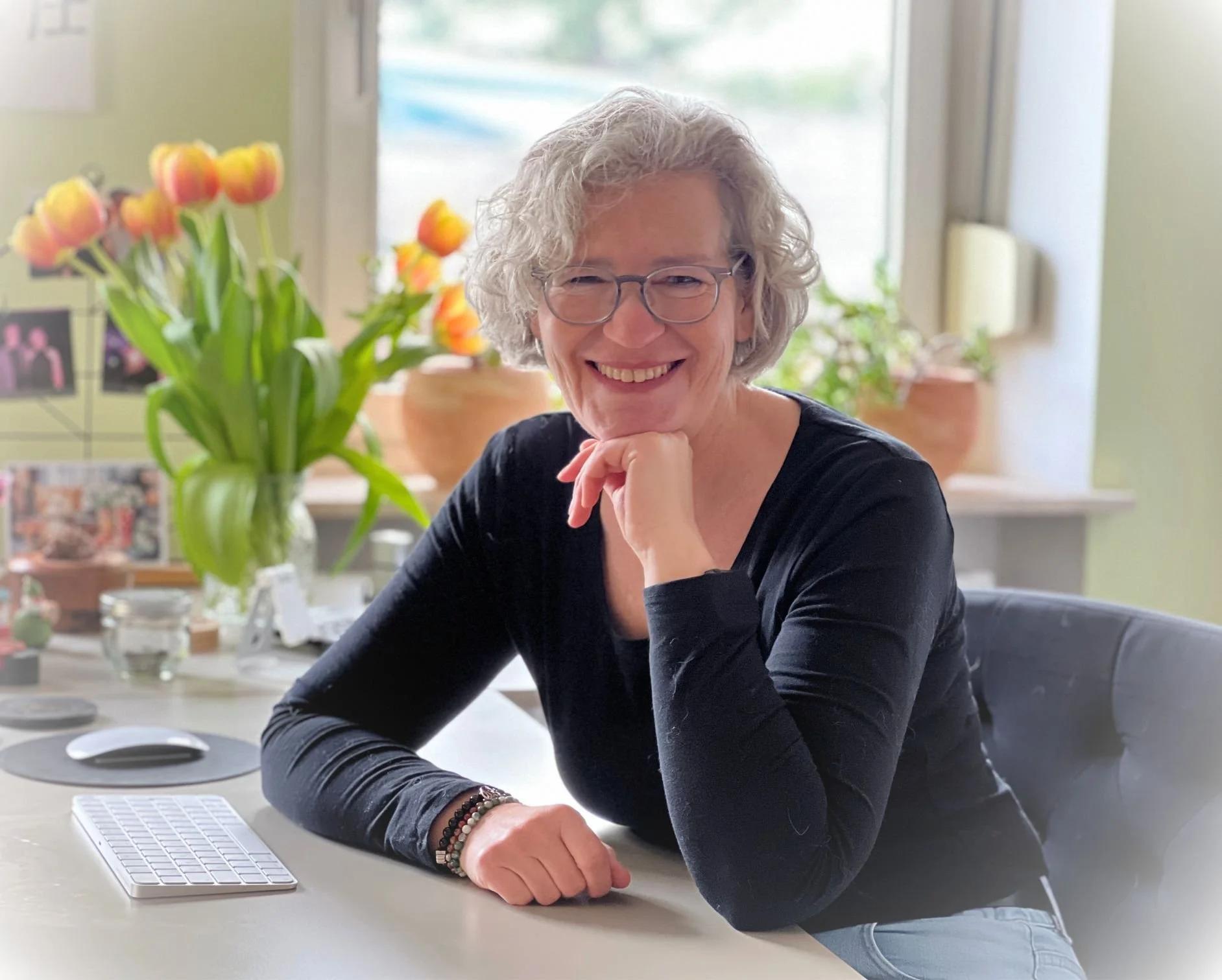 Eine lächelnde Frau mit grauen, lockigen Haaren vor einem Fenster mit Topfpflanzen und Tulpen, sitzend an einem Schreibtisch mit einer Computermaus und einer kleinen Tastatur.