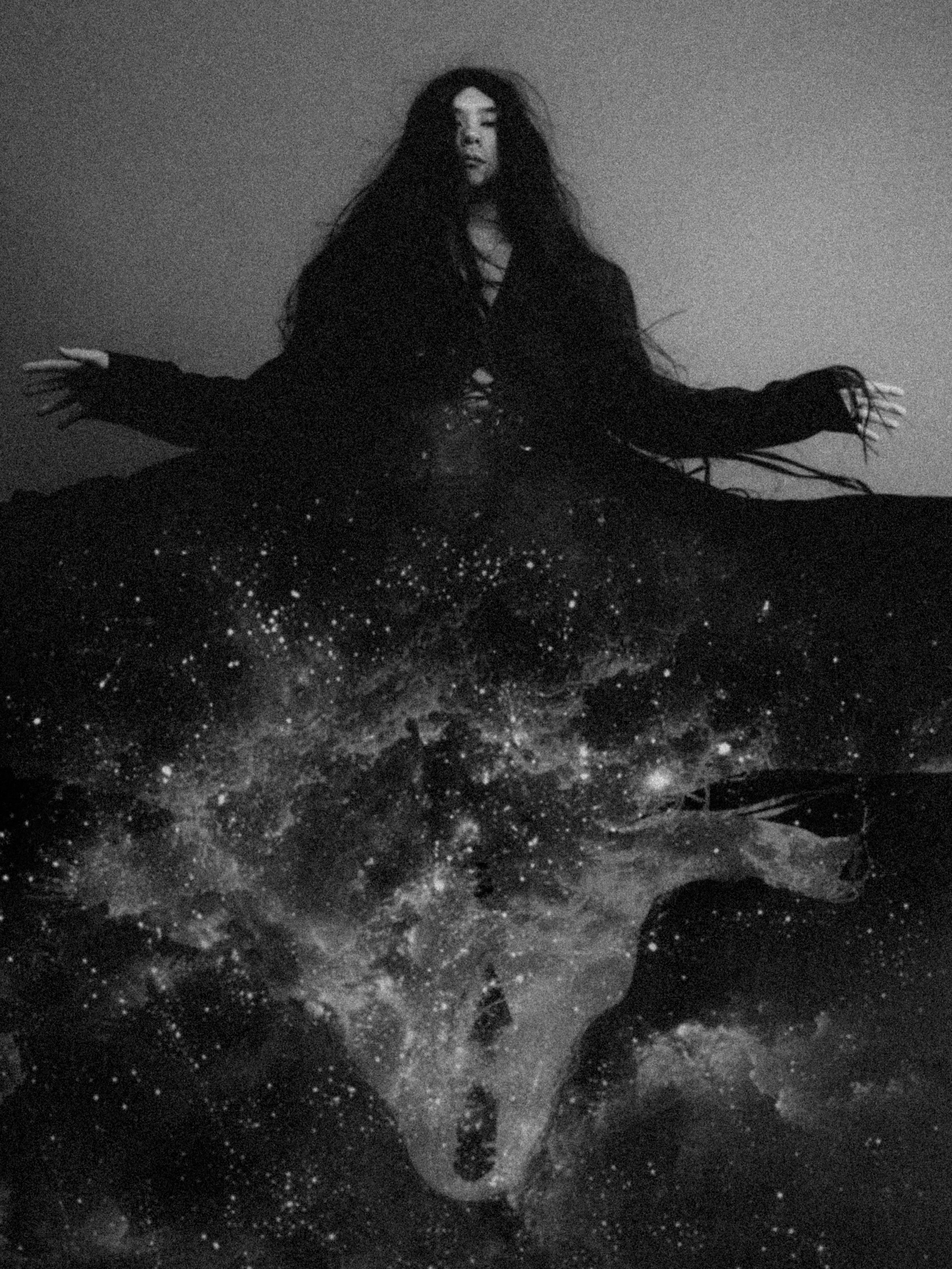 A woman with long dark hair wearing a dark dress with lace-up front, sitting on rocky terrain at night, surrounded by stars or sparkling lights.
