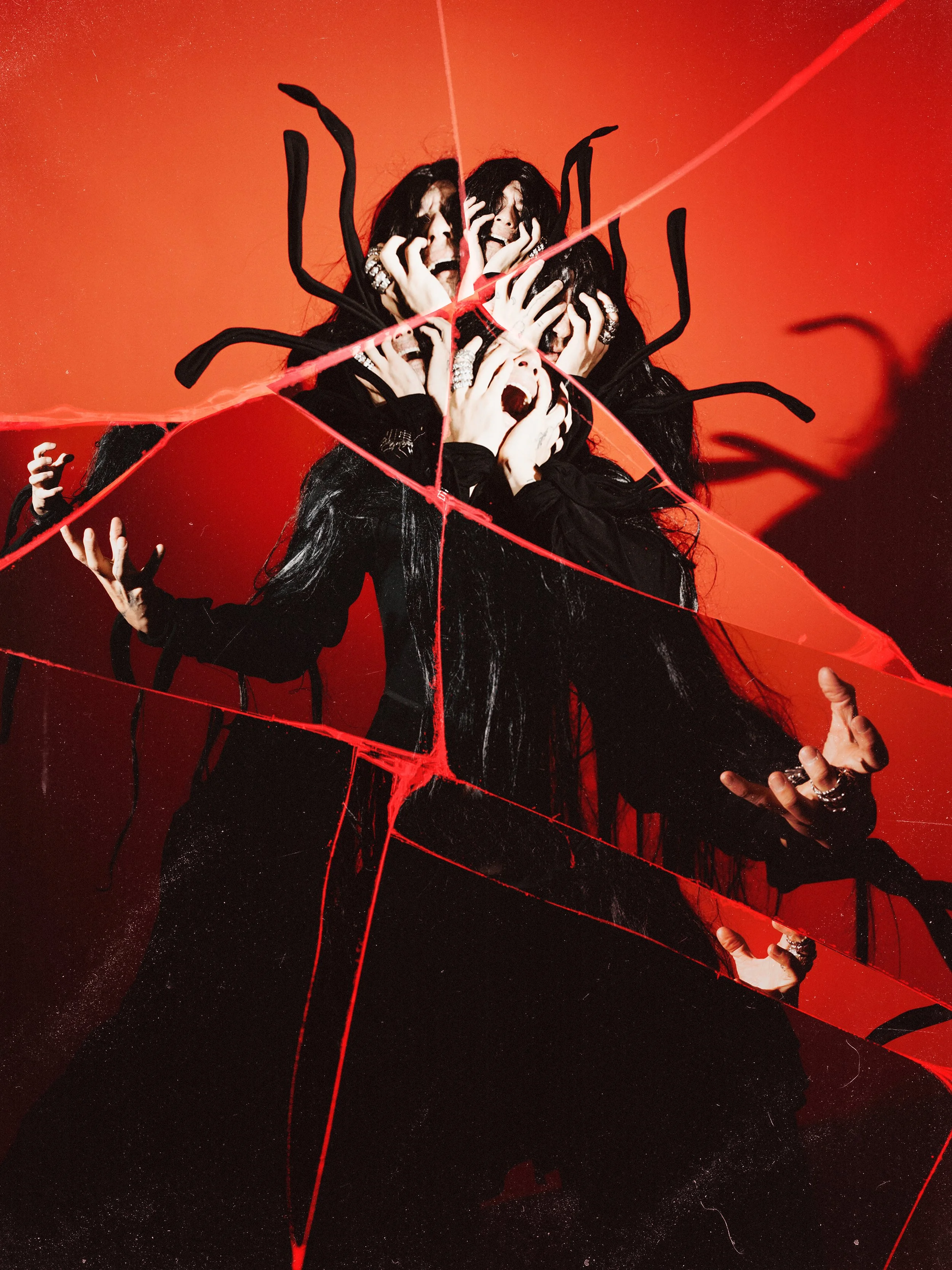 A person dressed in black with long black hair, multiple rings, and dark makeup, is seen behind a broken mirror with red lines and spider-like black decorations. The person appears to be screaming or showing a frightened expression against a vibrant red background.
