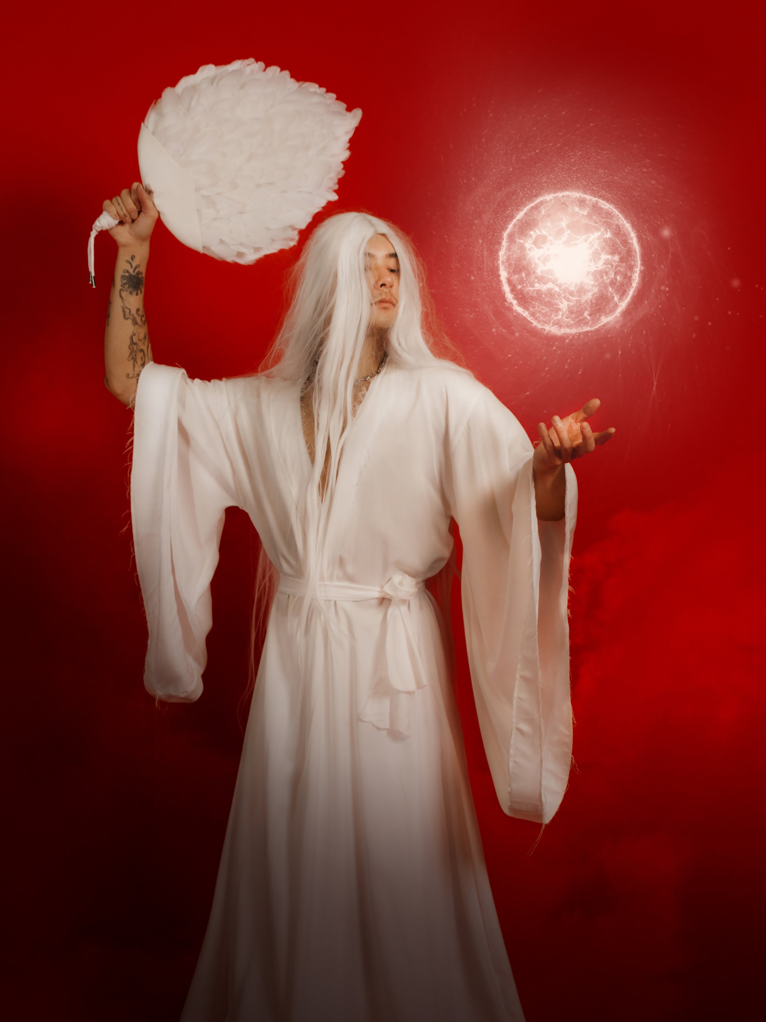 Person dressed as an angel with long white hair, wearing a white robe, holding a feathered fan, standing against a red background with a glowing celestial body.