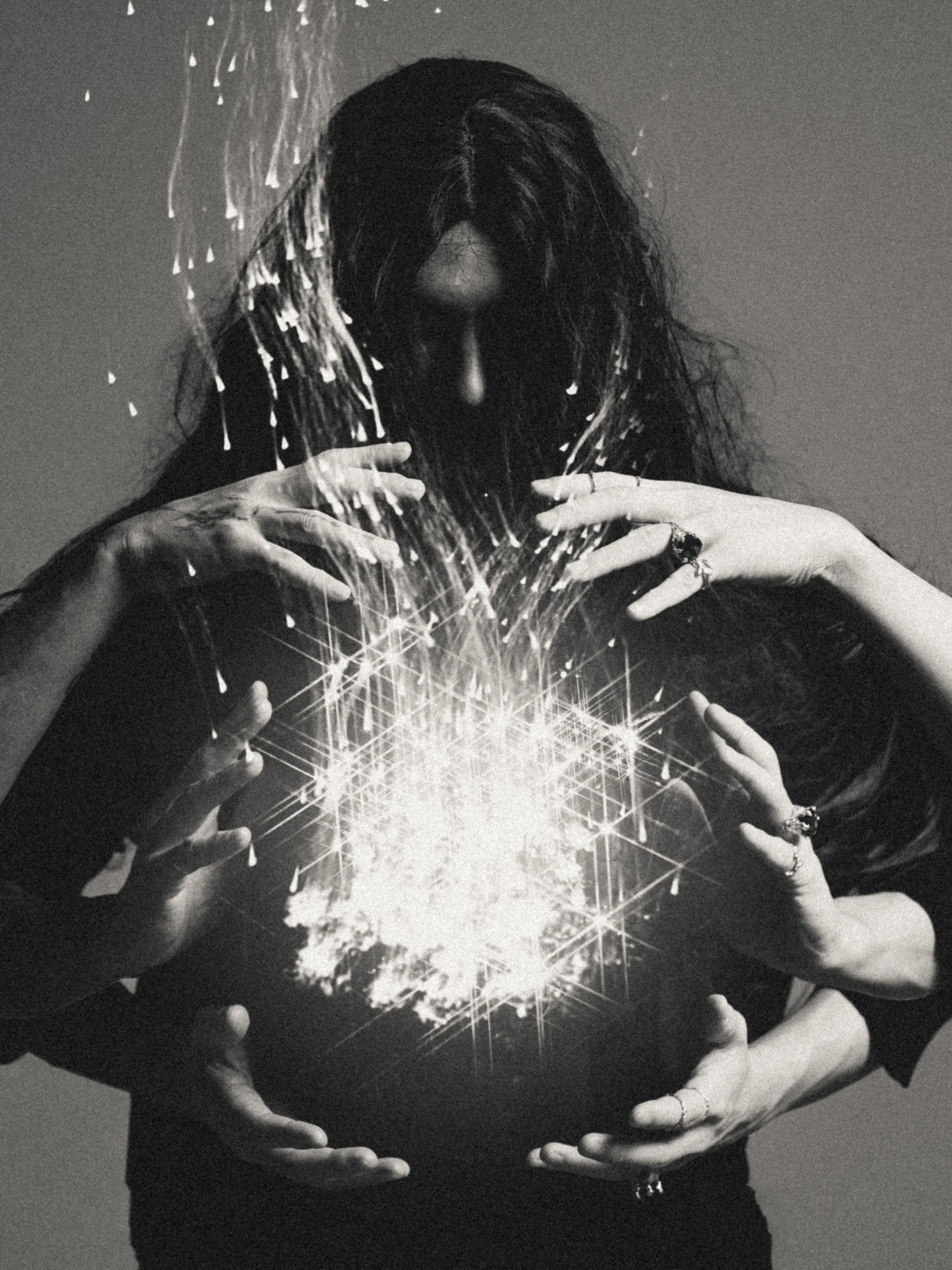 A person with long dark hair, wearing rings and bracelets, holding a sphere emitting bright light with star-like sparkles and geometric patterns.