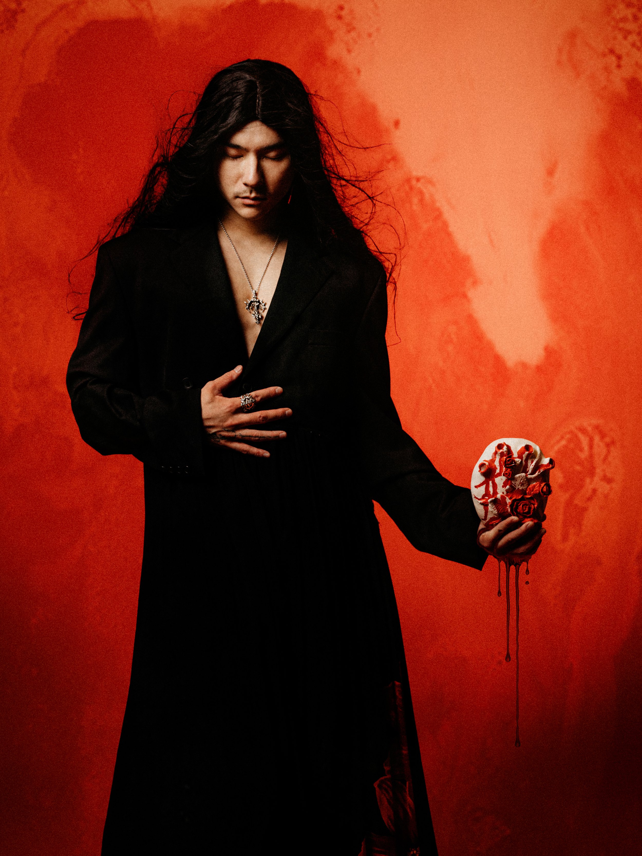 A person with long dark hair and a black blazer holding a heart-shaped cake with red roses, against an orange-red background.