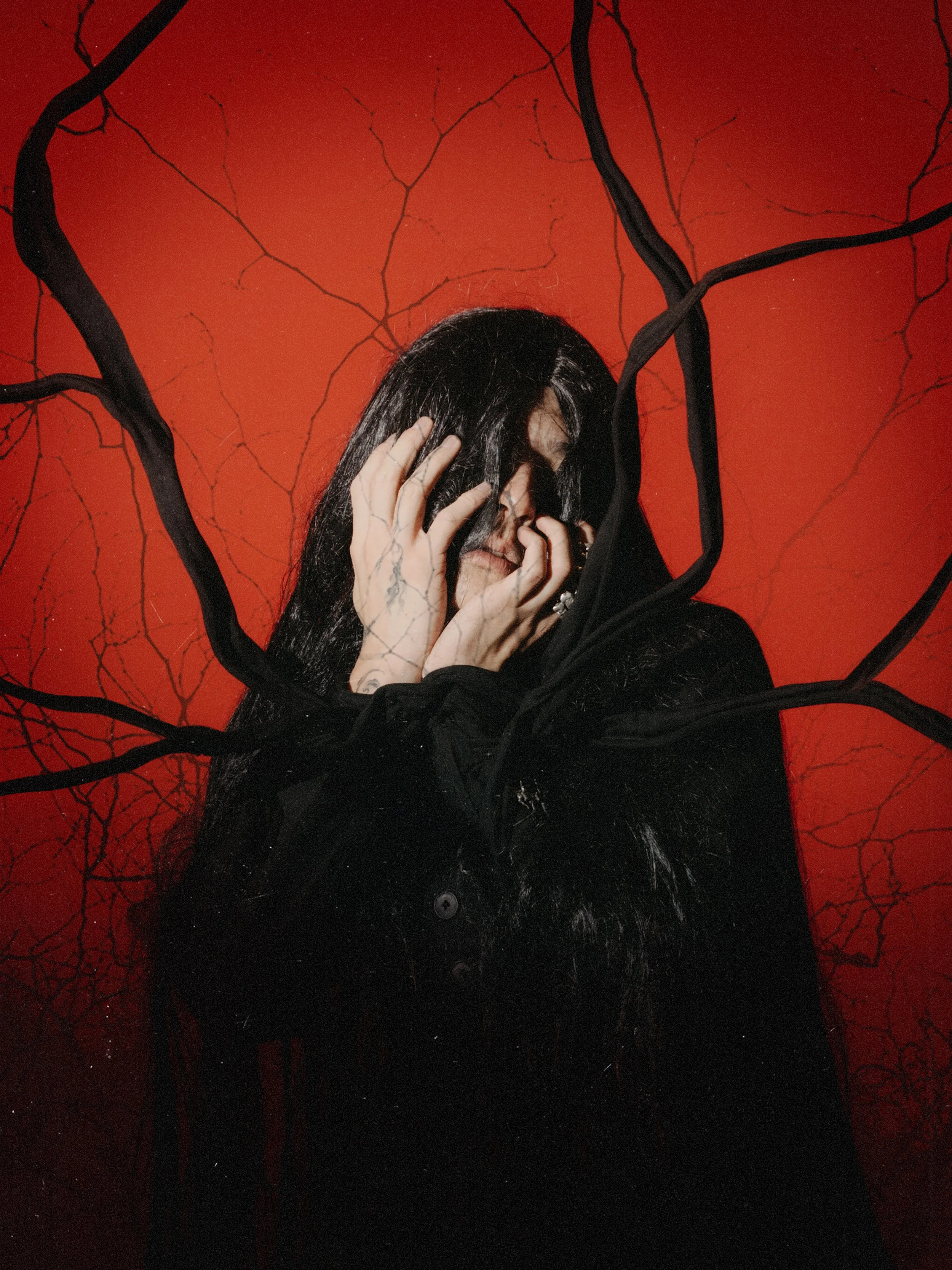 A person with long black hair and tattoos on their hand is holding their face with both hands, covering part of their face, against a red background with black, tree-branch-like designs.