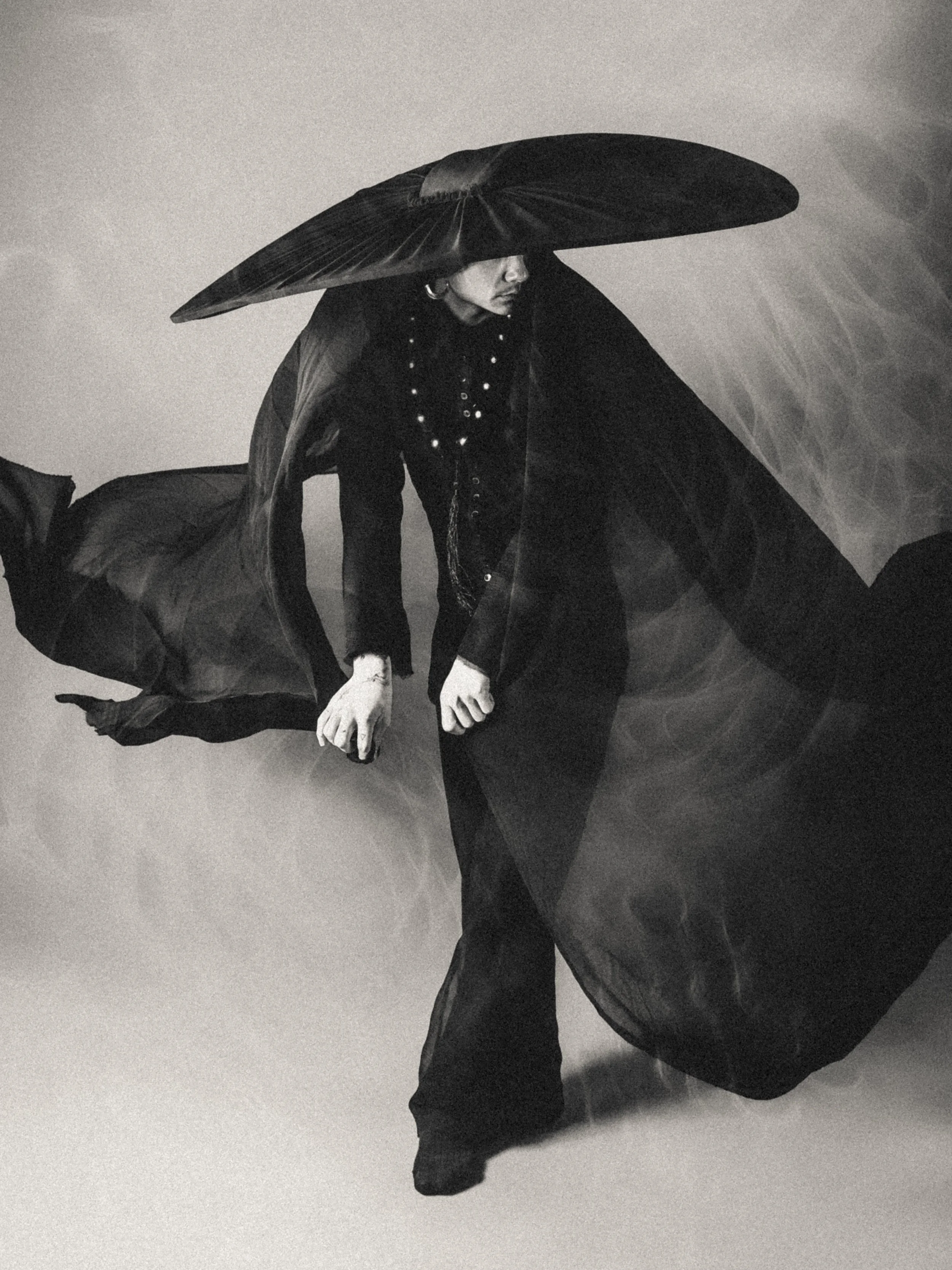 A person crouching with an oversized leaf covering their head, dressed in dark clothing, in black and white.