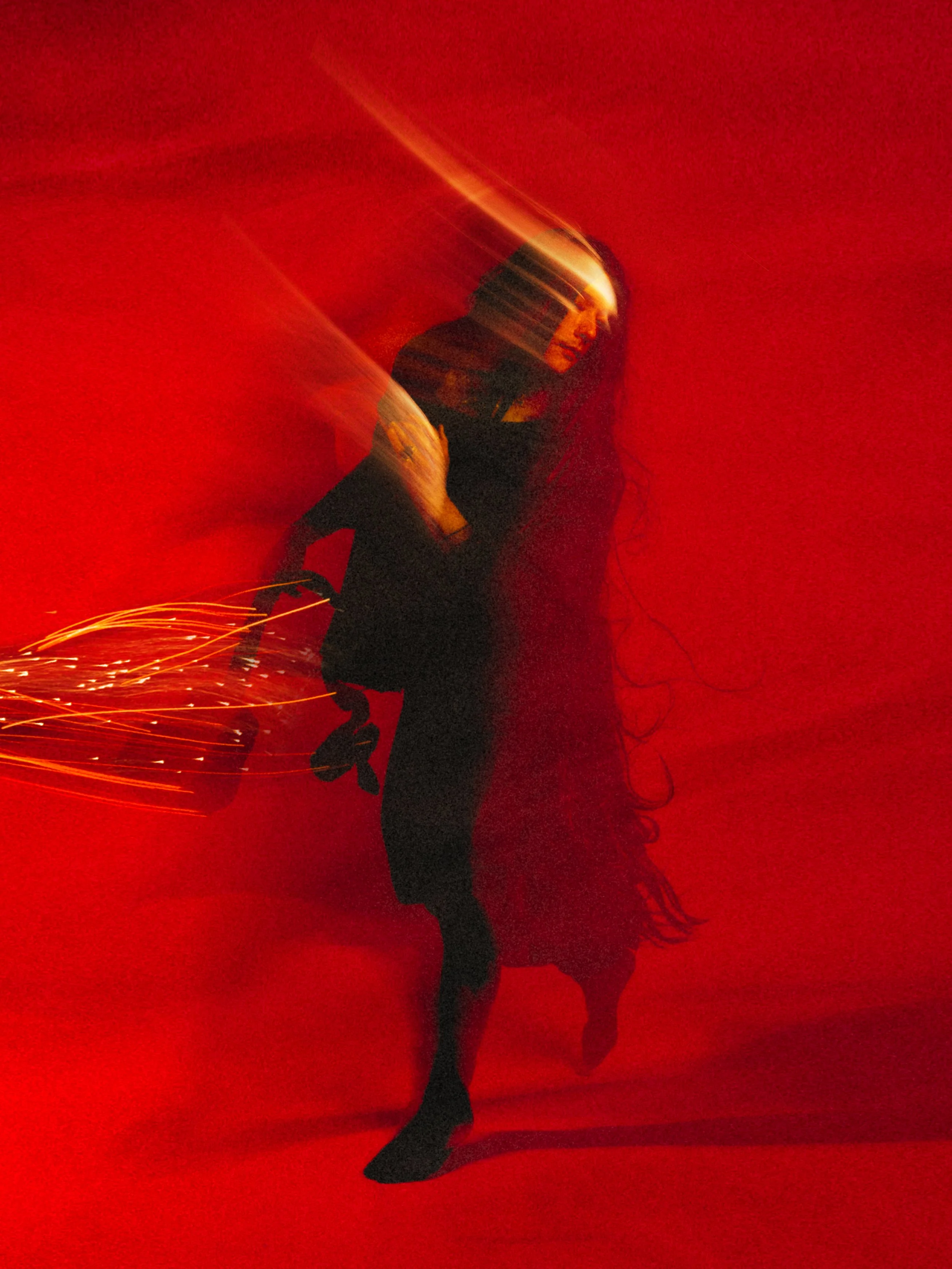 A woman with long hair in black clothing performing a dance or pose against a red background, with light trails and motion blur.